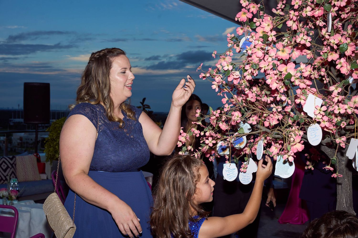 Women decorating a pink cherry blossom tree with ornaments at an outdoor evening event.