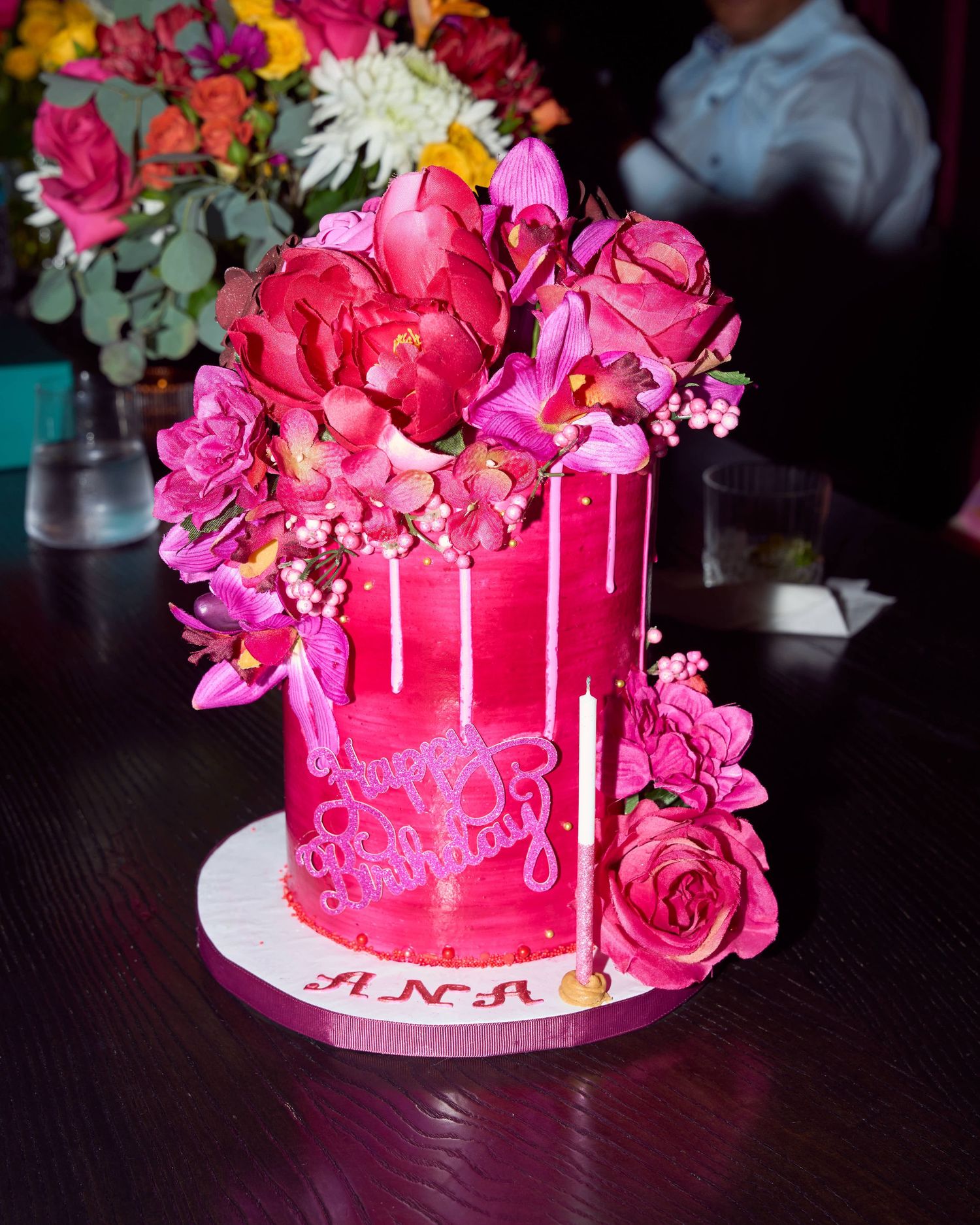 Elegant pink birthday cake with floral decorations and drip frosting, reading Happy Birthday ANA.