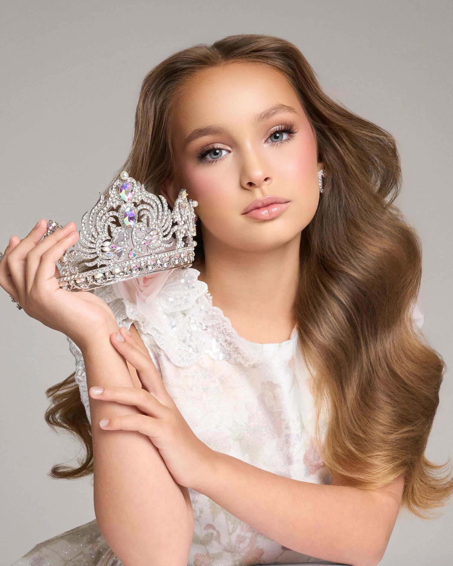 Young beauty queen holds sparkling tiara crown in elegant white lace dress with wavy hair.