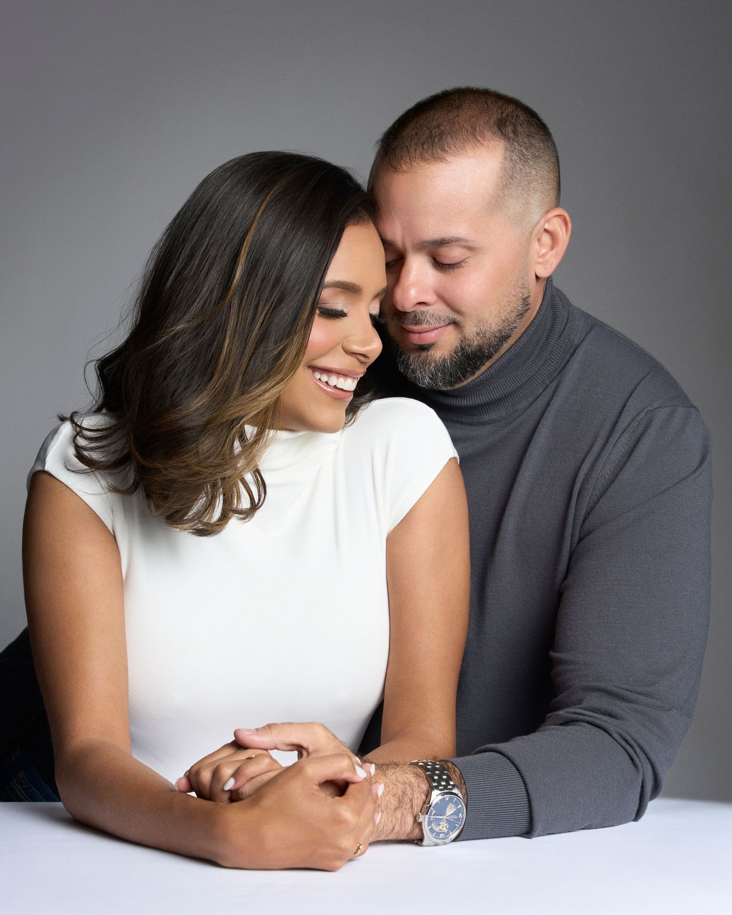 Smiling couple sharing a tender moment, man embracing woman from behind while holding hands at a studio portrait session.