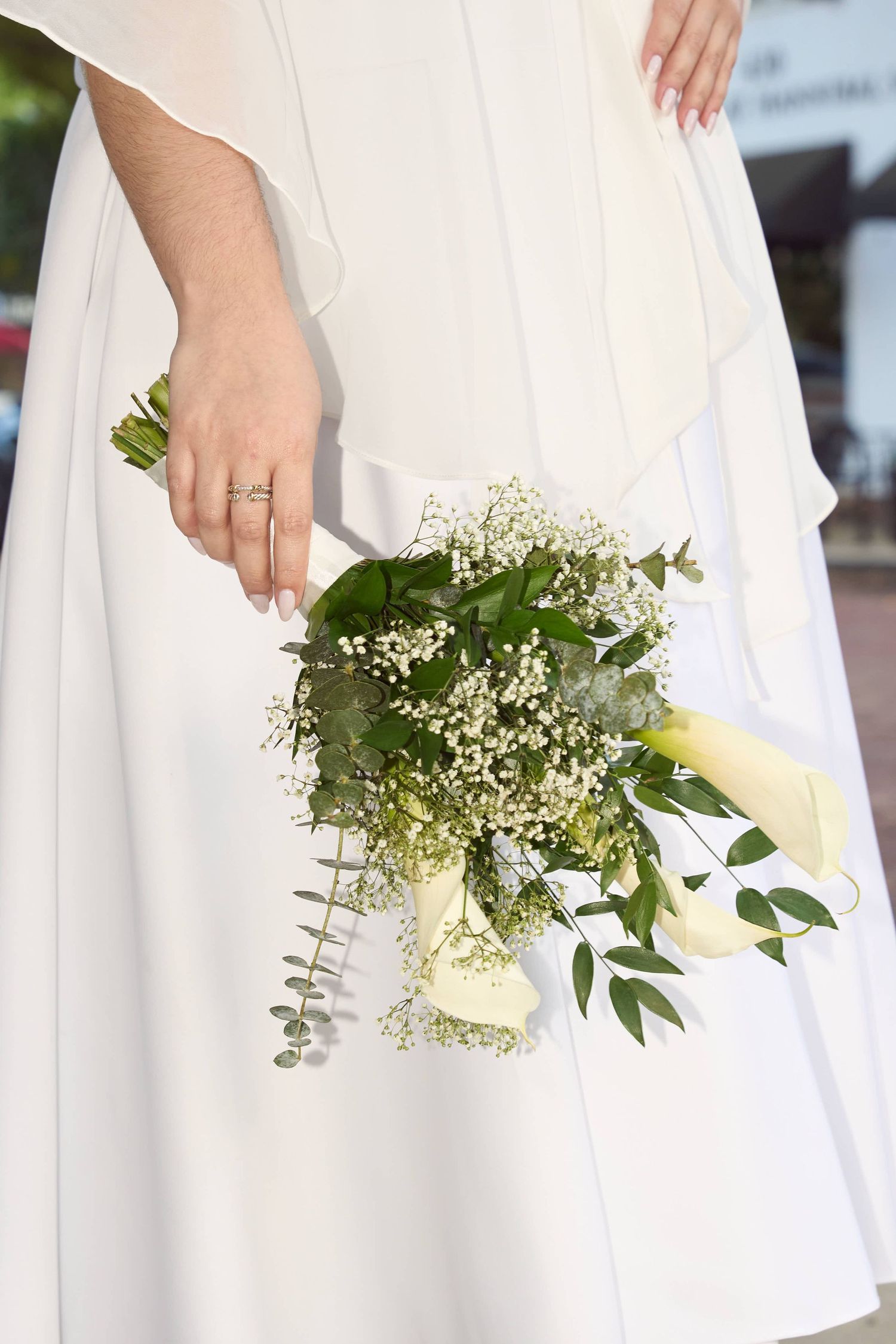 Bride in white dress holding elegant bouquet of white roses and greenery with wedding ring.