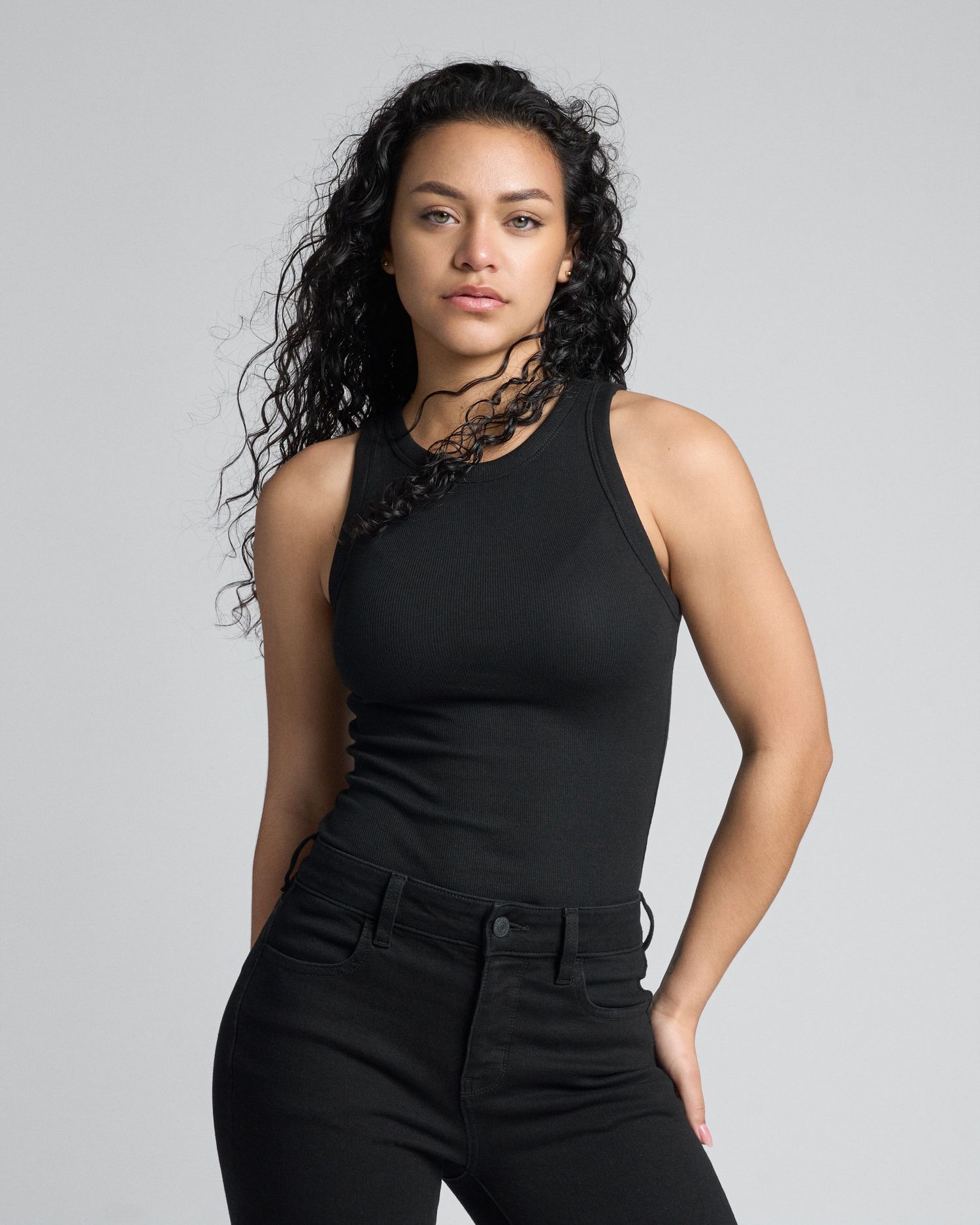 Woman with curly black hair wearing black sleeveless bodysuit and black pants in studio.