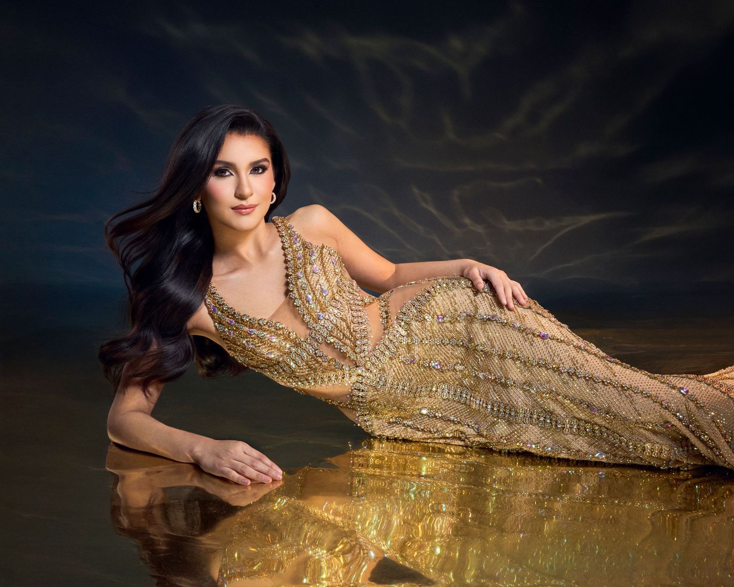 Elegant woman in gold beaded gown poses reclined against reflective golden surface.