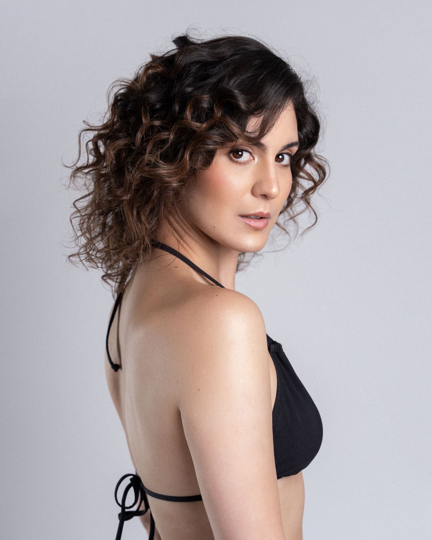 Close-up of a woman with curly dark hair wearing a black halter bikini top in a studio setting.