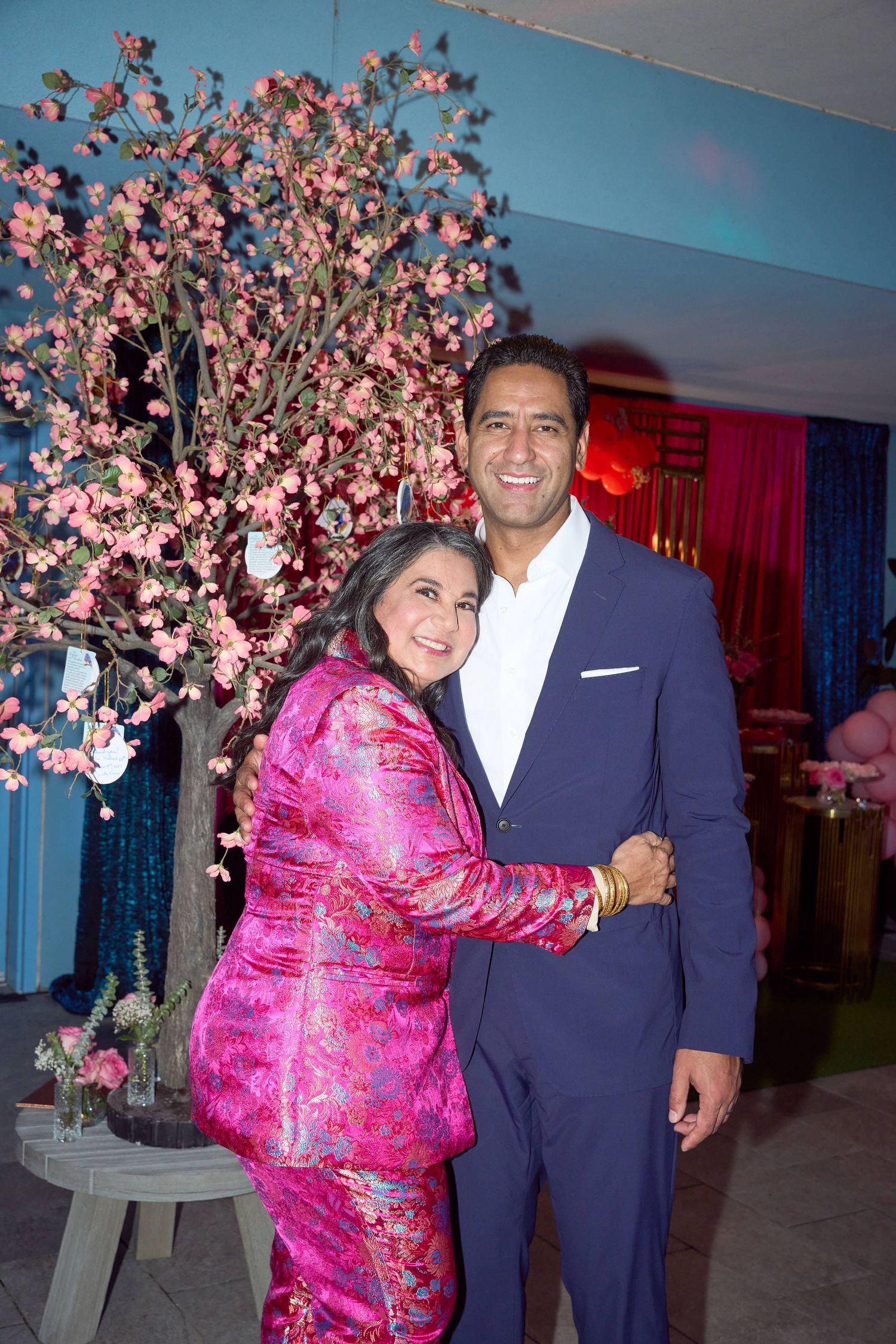A woman in pink floral suit and man in navy blue suit embrace smiling near a cherry blossom tree at an event.