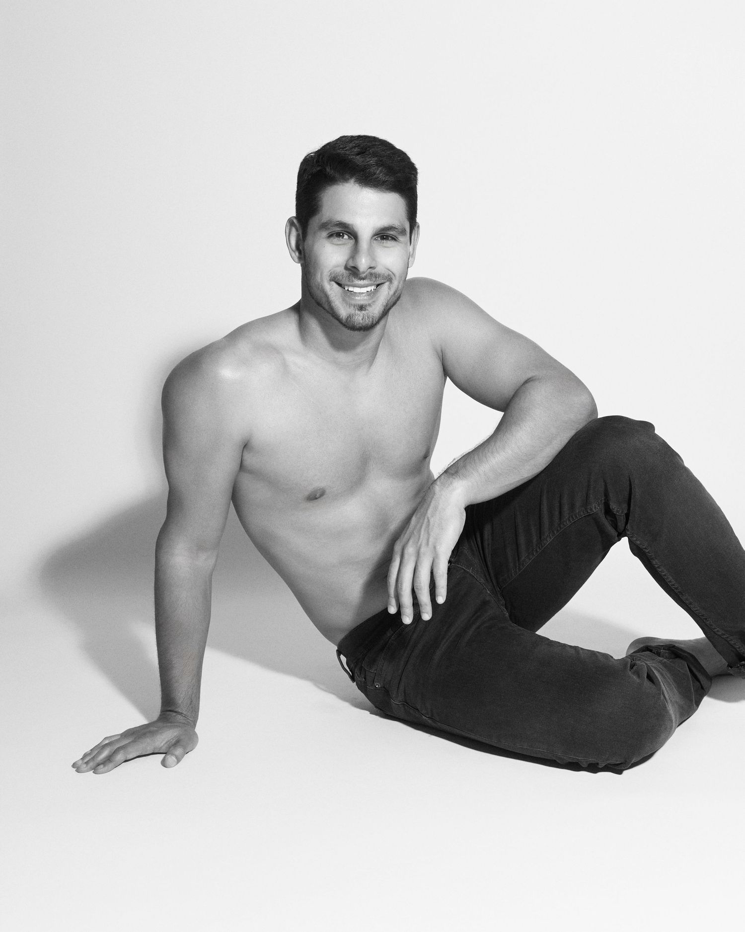 Black and white photo of a shirtless muscular man sitting on floor, smiling, wearing dark jeans.