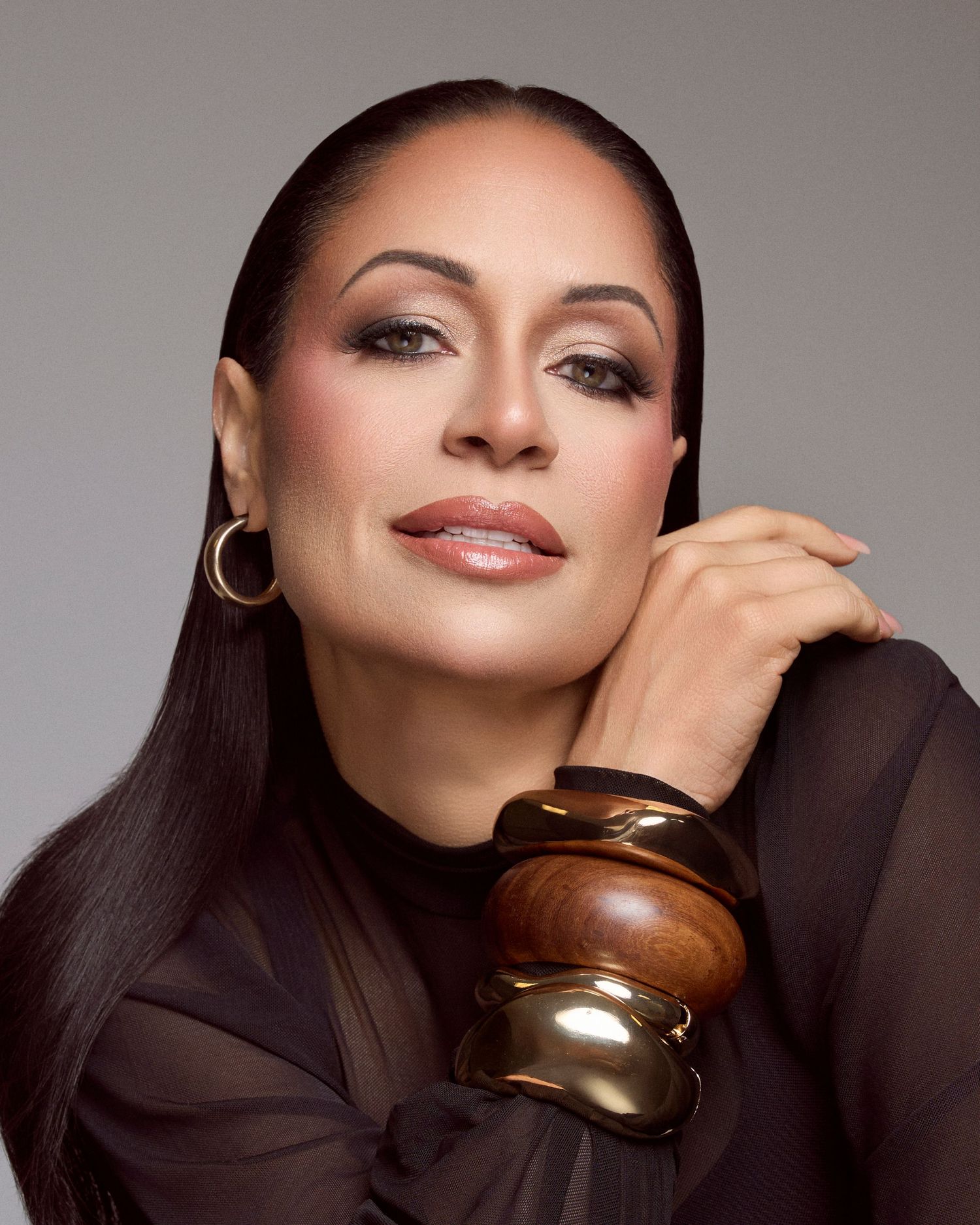 Elegant woman in black sheer top with gold and wood bangles posing gracefully against gray background.