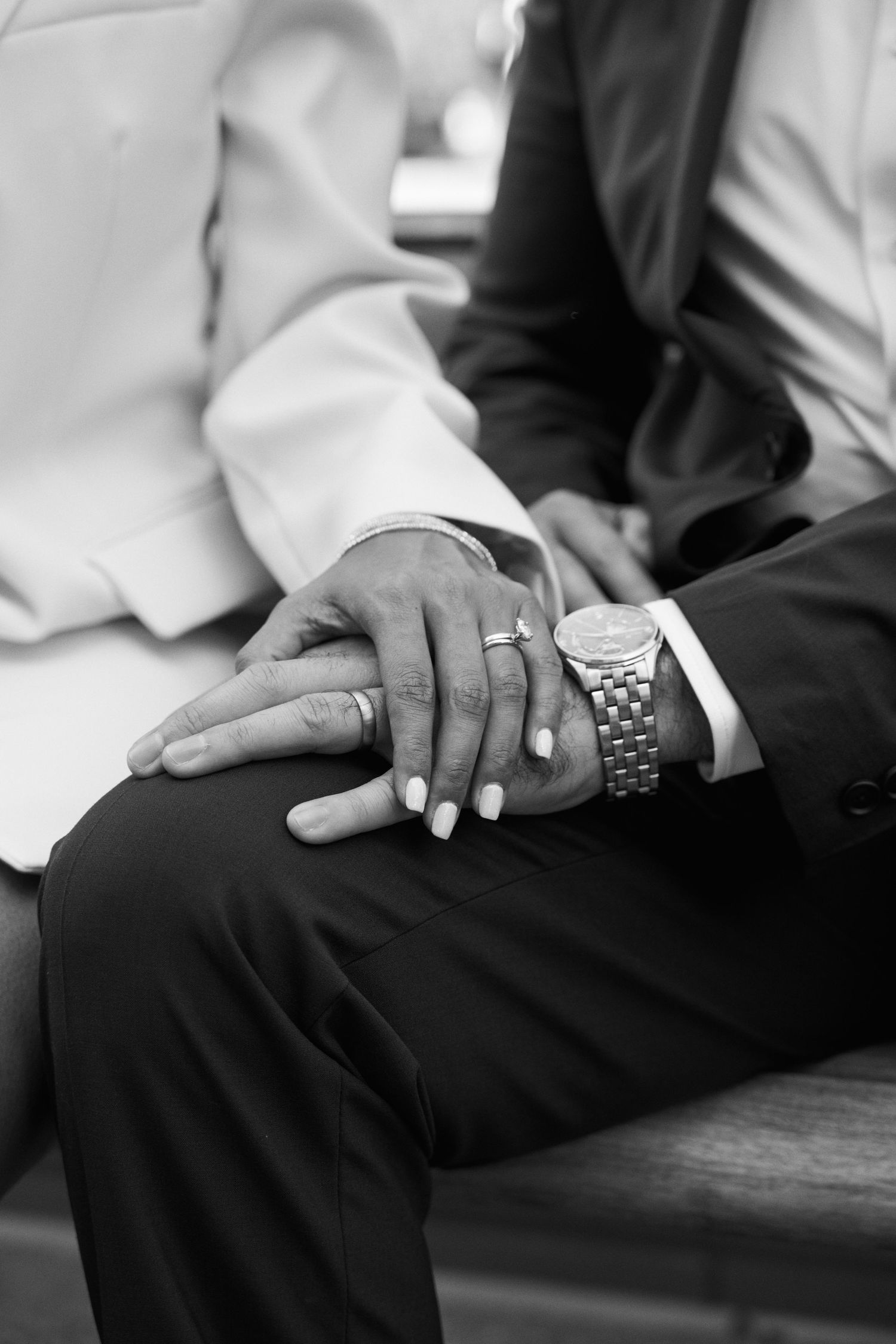 Black and white close-up of newlyweds holding hands showing wedding rings and watch on bench.