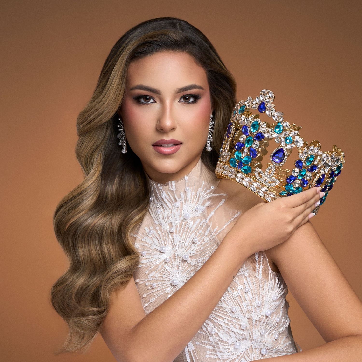 Beauty queen holds ornate blue and gold crown wearing white embellished high-neck gown.
