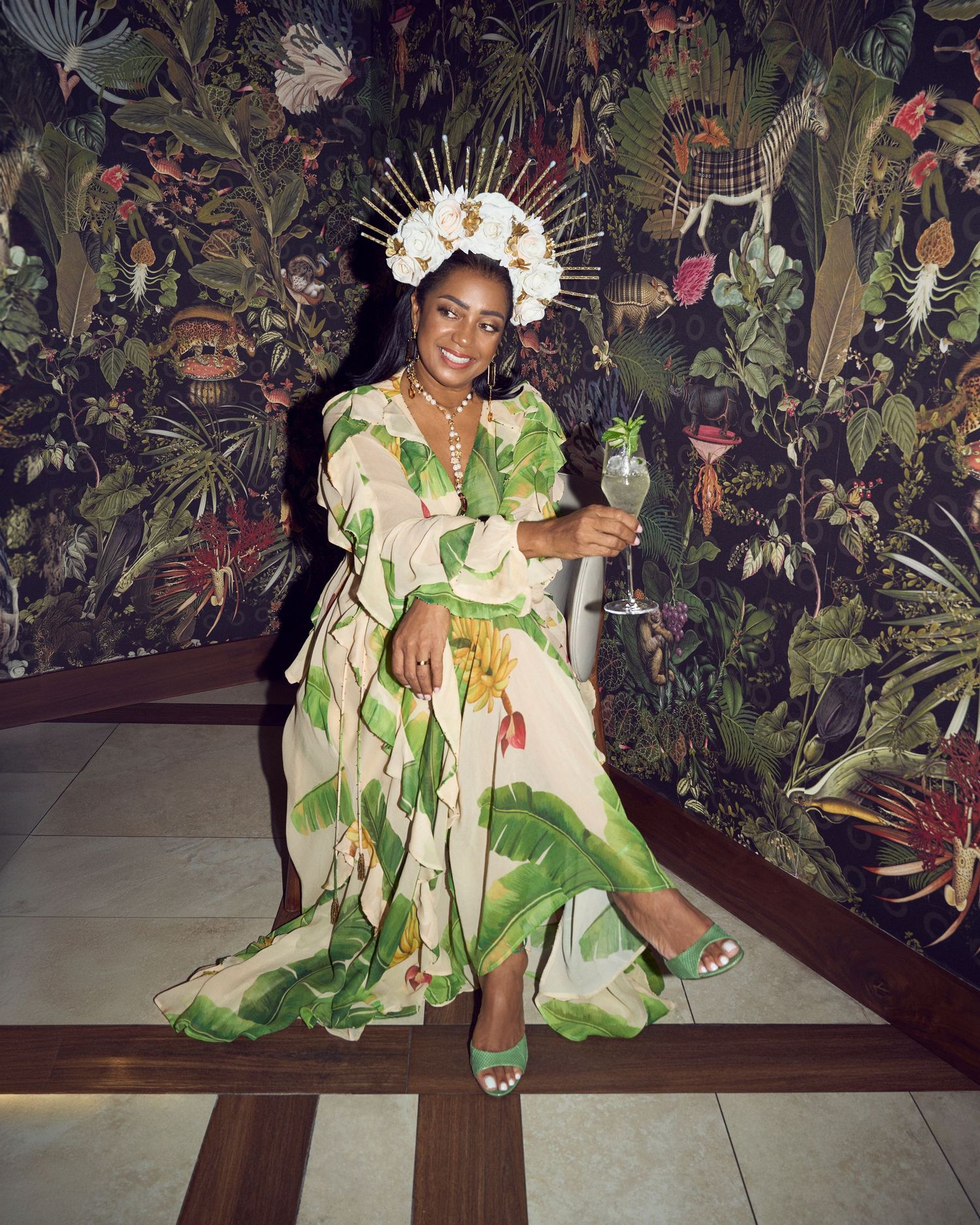 Woman in floral green dress and sunburst crown holds wine glass, posing before lush tropical botanical wallpaper.
