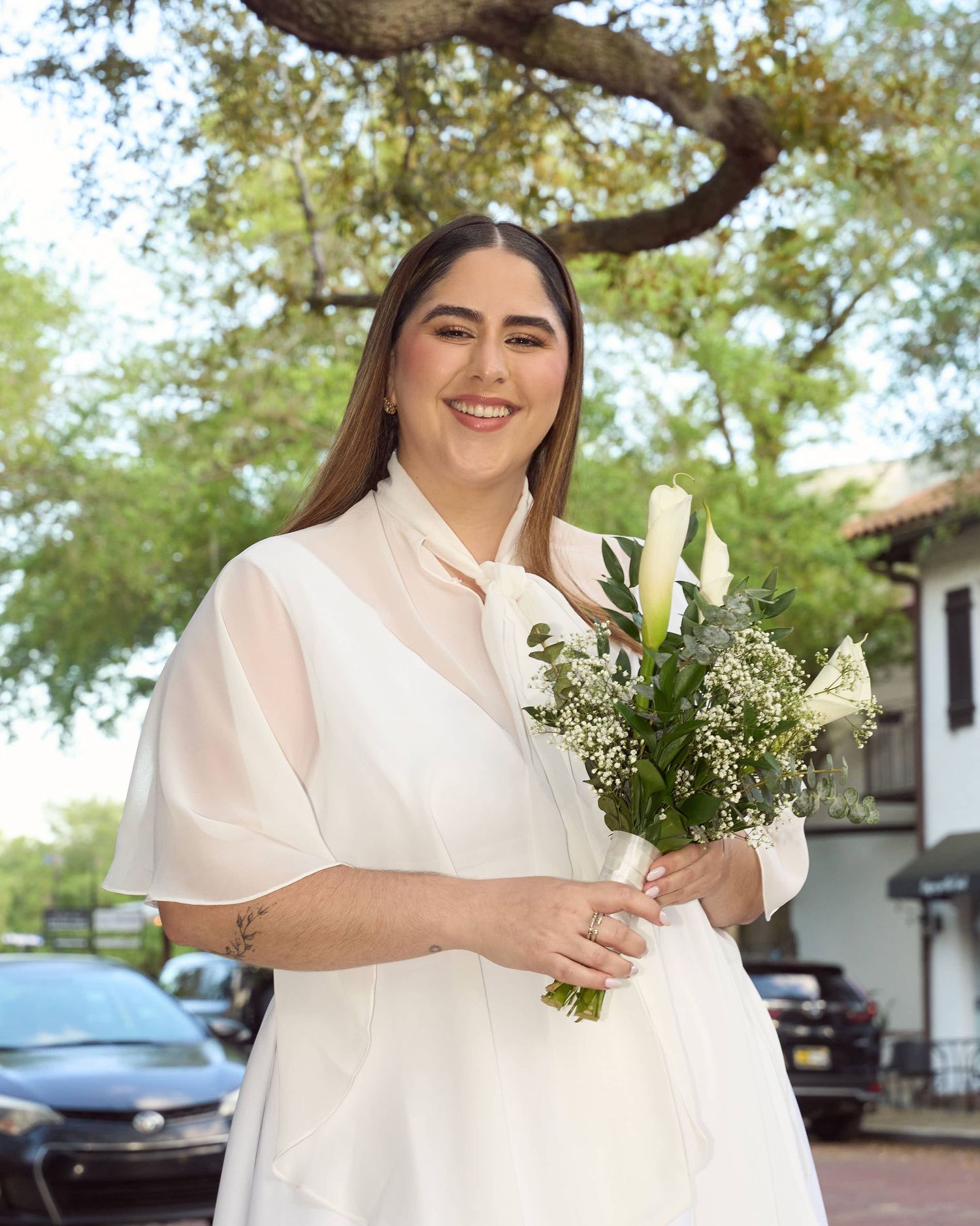 Smiling bride in white dress holding calla lily bouquet outdoors under green trees.
