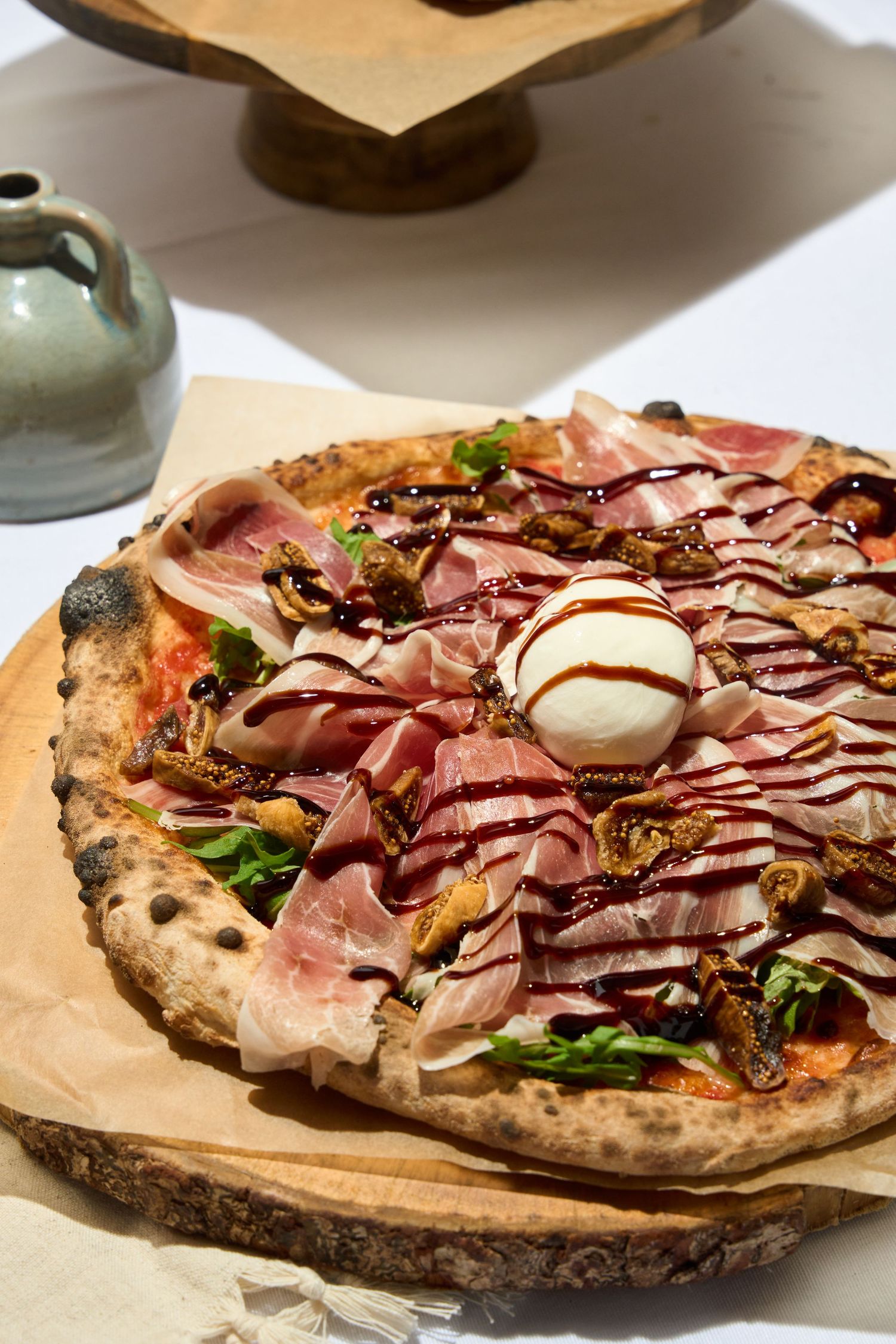 Neapolitan pizza topped with prosciutto, burrata, figs, arugula, and balsamic glaze on wooden board.