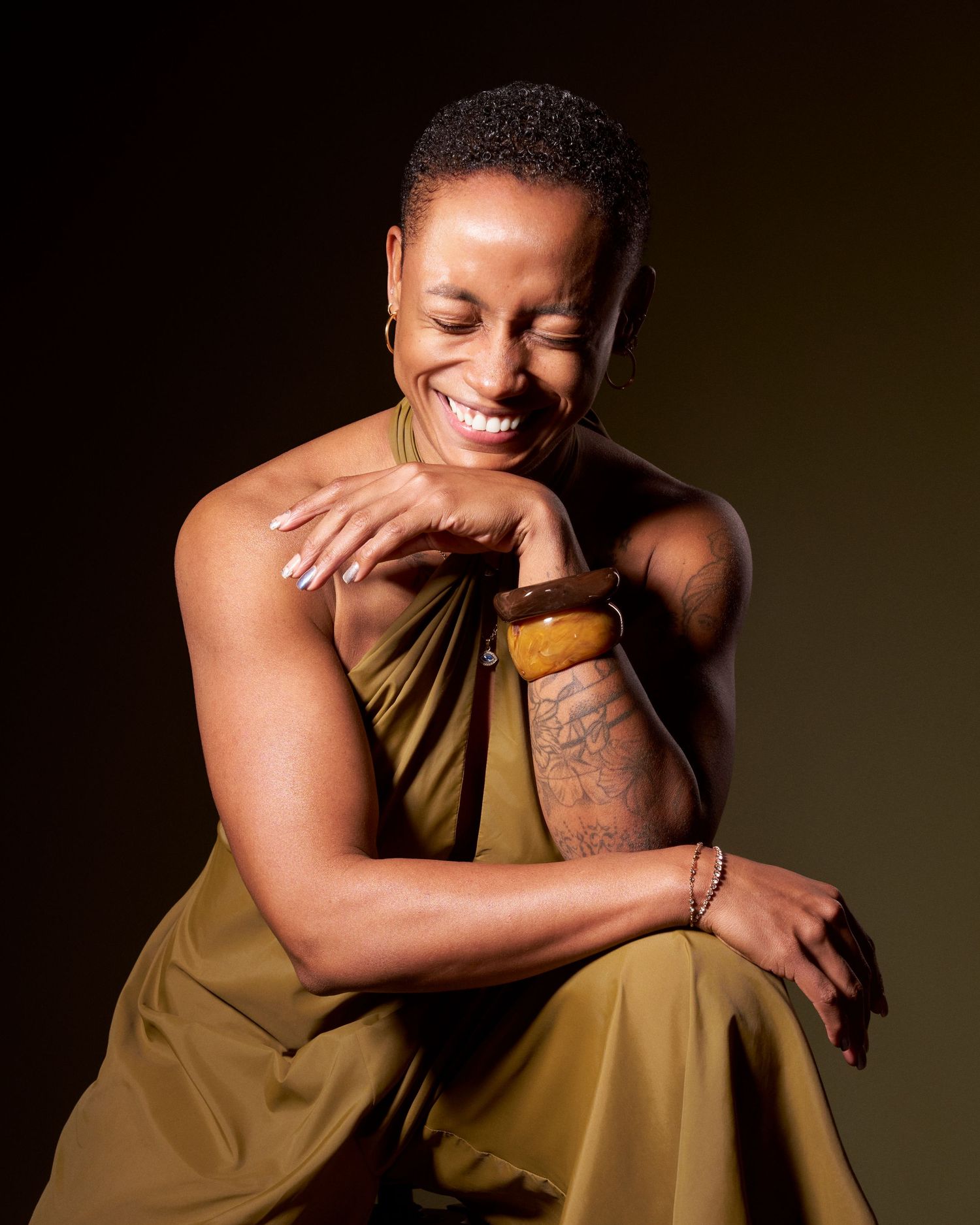 Woman in olive dress laughs joyfully while seated against dark background.
