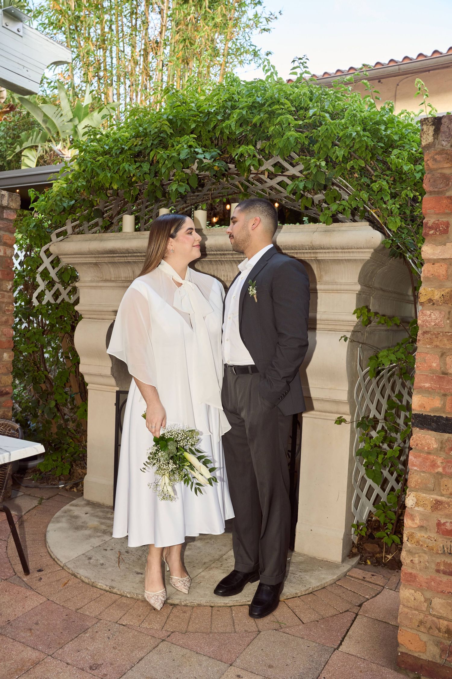 Wedding couple gazing at each other under ivy-covered arch at outdoor garden ceremony.