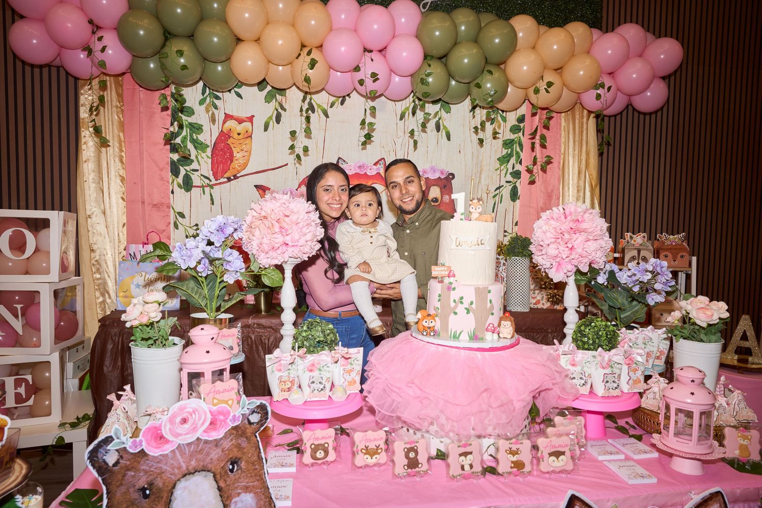 Family posing at a pink owl-themed first birthday party with balloon arch, floral arrangements, and decorated cake table.