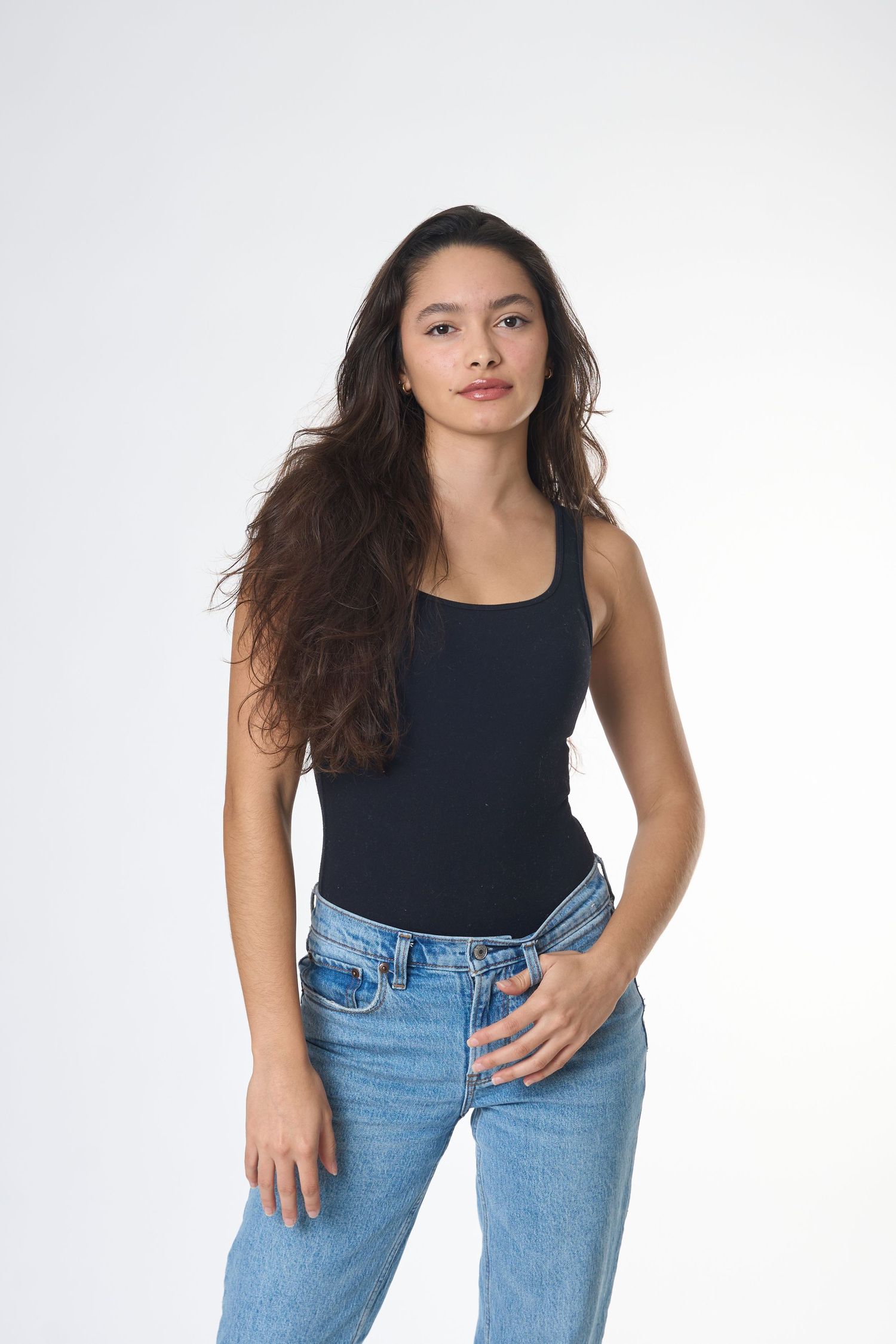 Young woman with long dark hair wearing a black tank top and blue jeans posing against a white background.