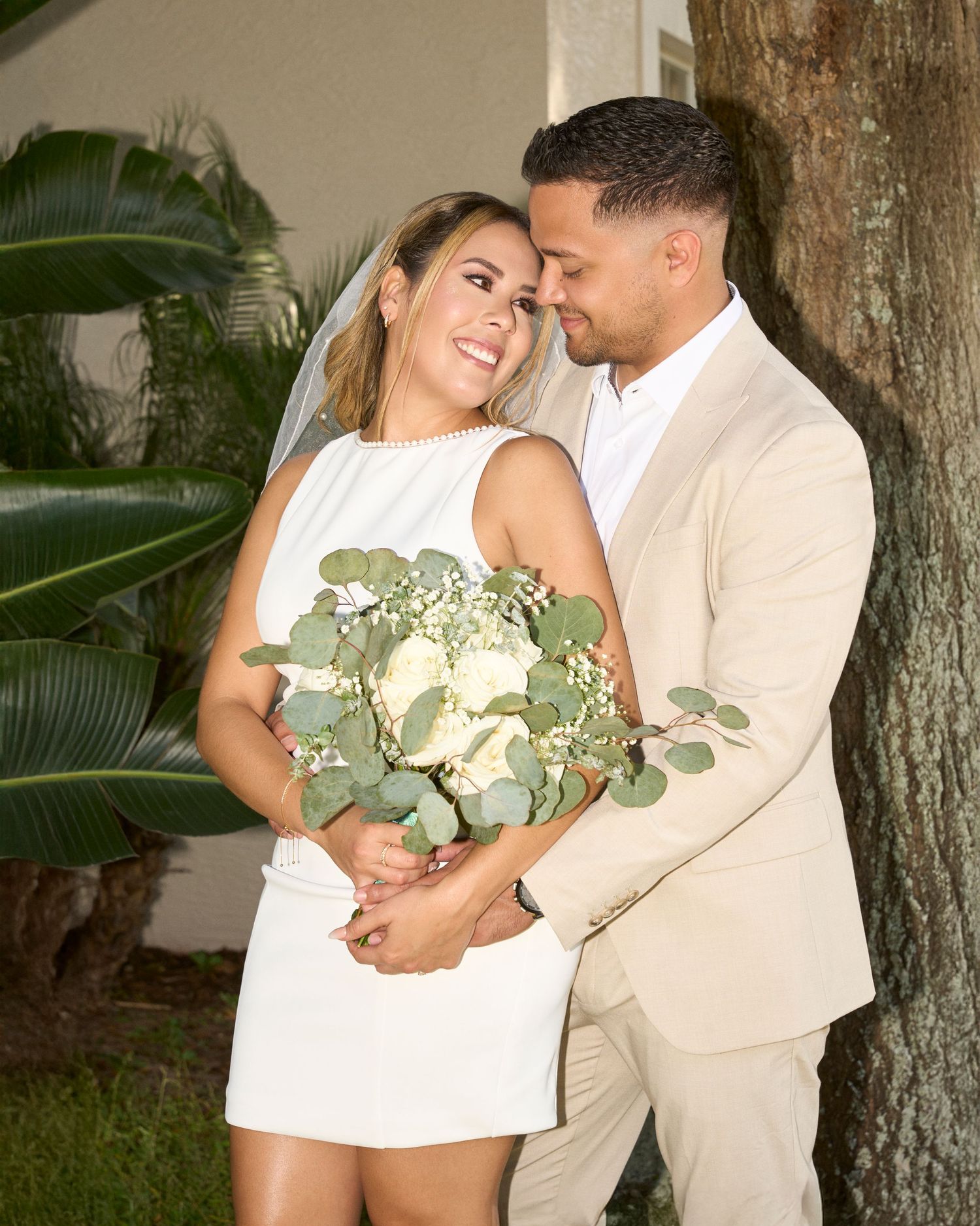 Bride in white mini dress holding bouquet embraced by groom in tan suit during outdoor wedding.