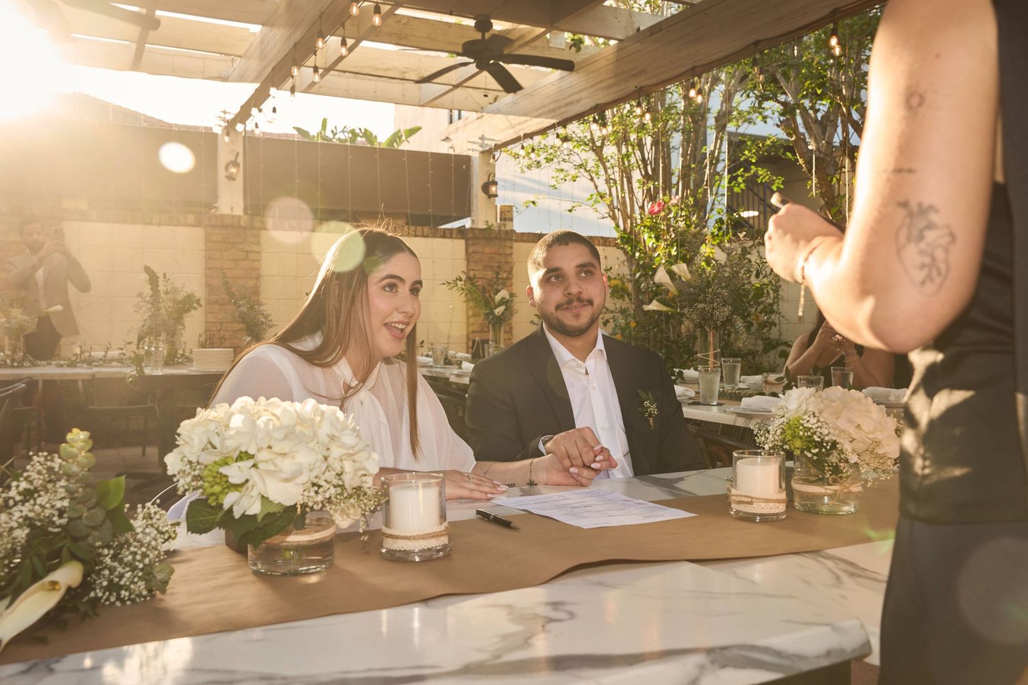 A smiling bride and groom sign their wedding documents at an outdoor reception table adorned with white floral centerpieces.