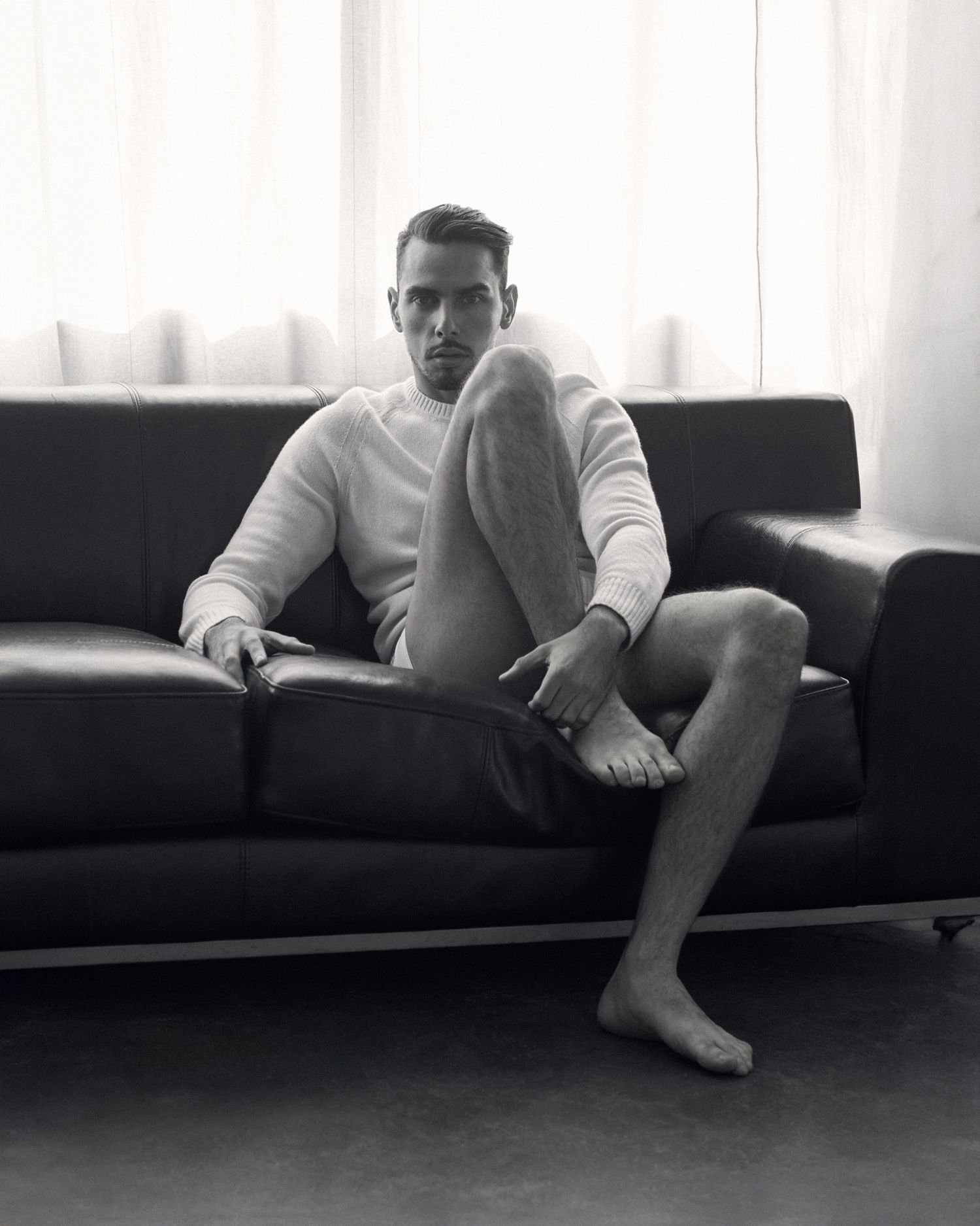 Black and white photo of a man in white sweater sitting barefoot on a leather sofa near a bright window.