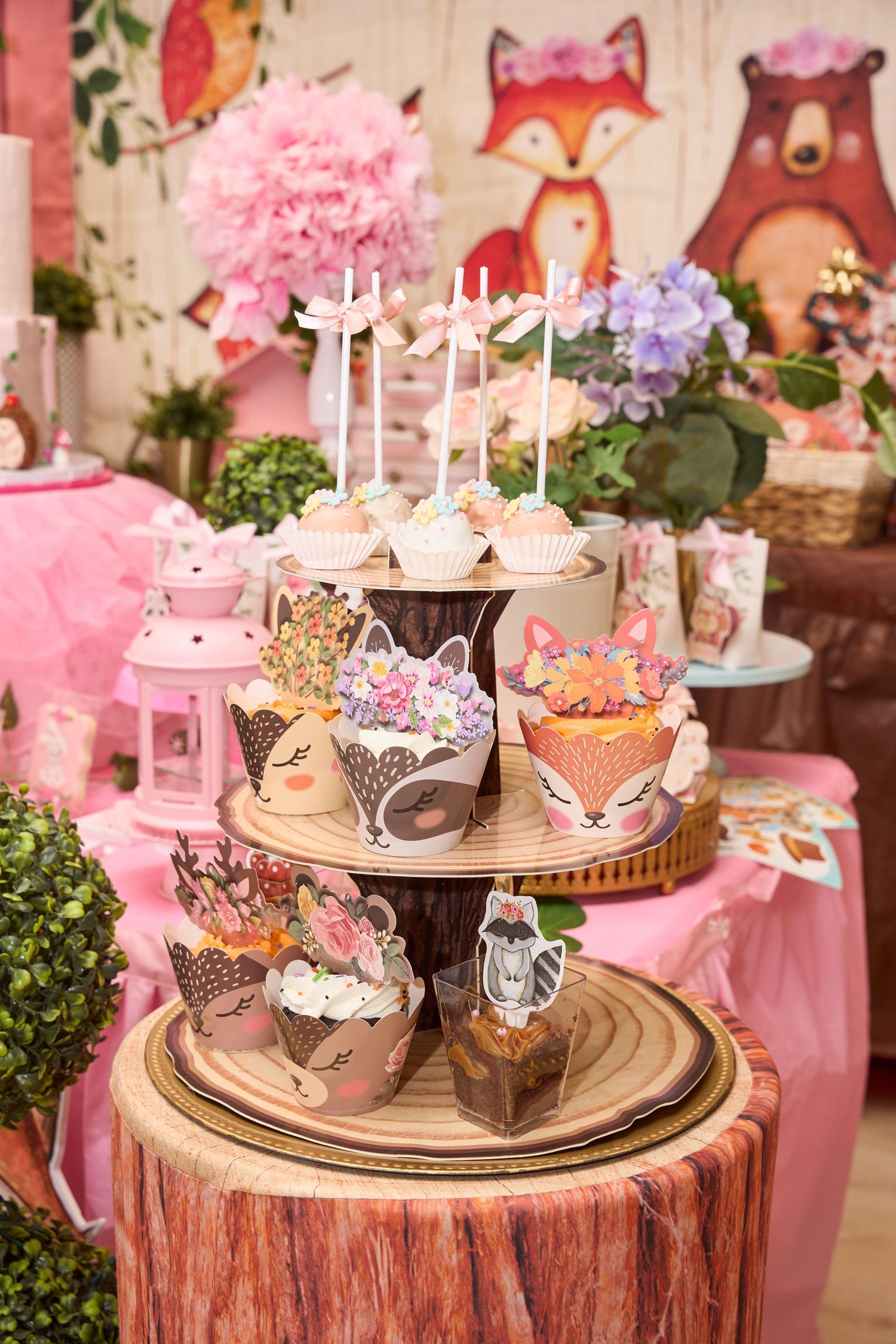 Woodland animal themed birthday party dessert table with fox and bear cupcakes on a rustic wood stand with pink decor.