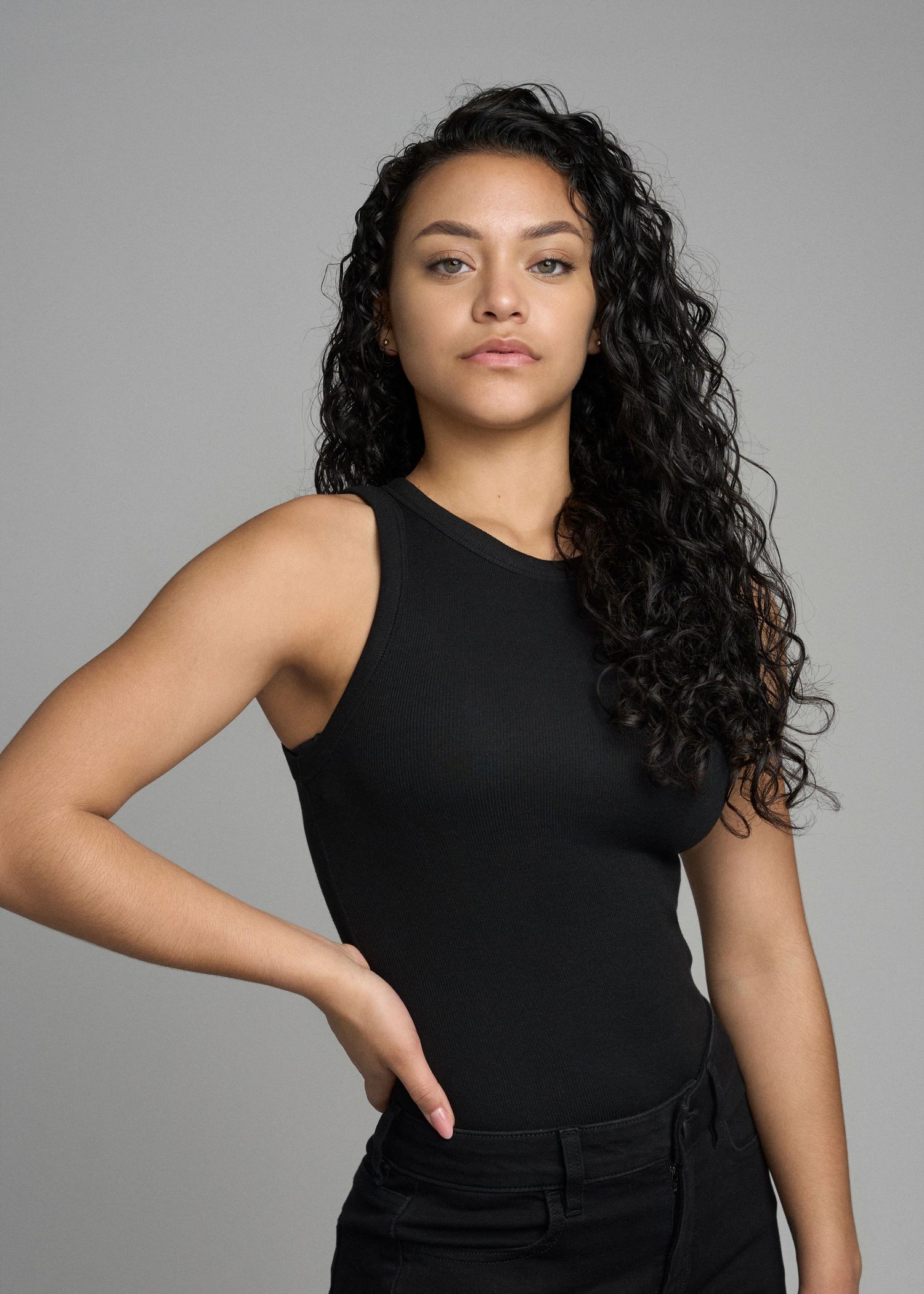 Woman with curly black hair posing in black sleeveless top and black pants in studio.