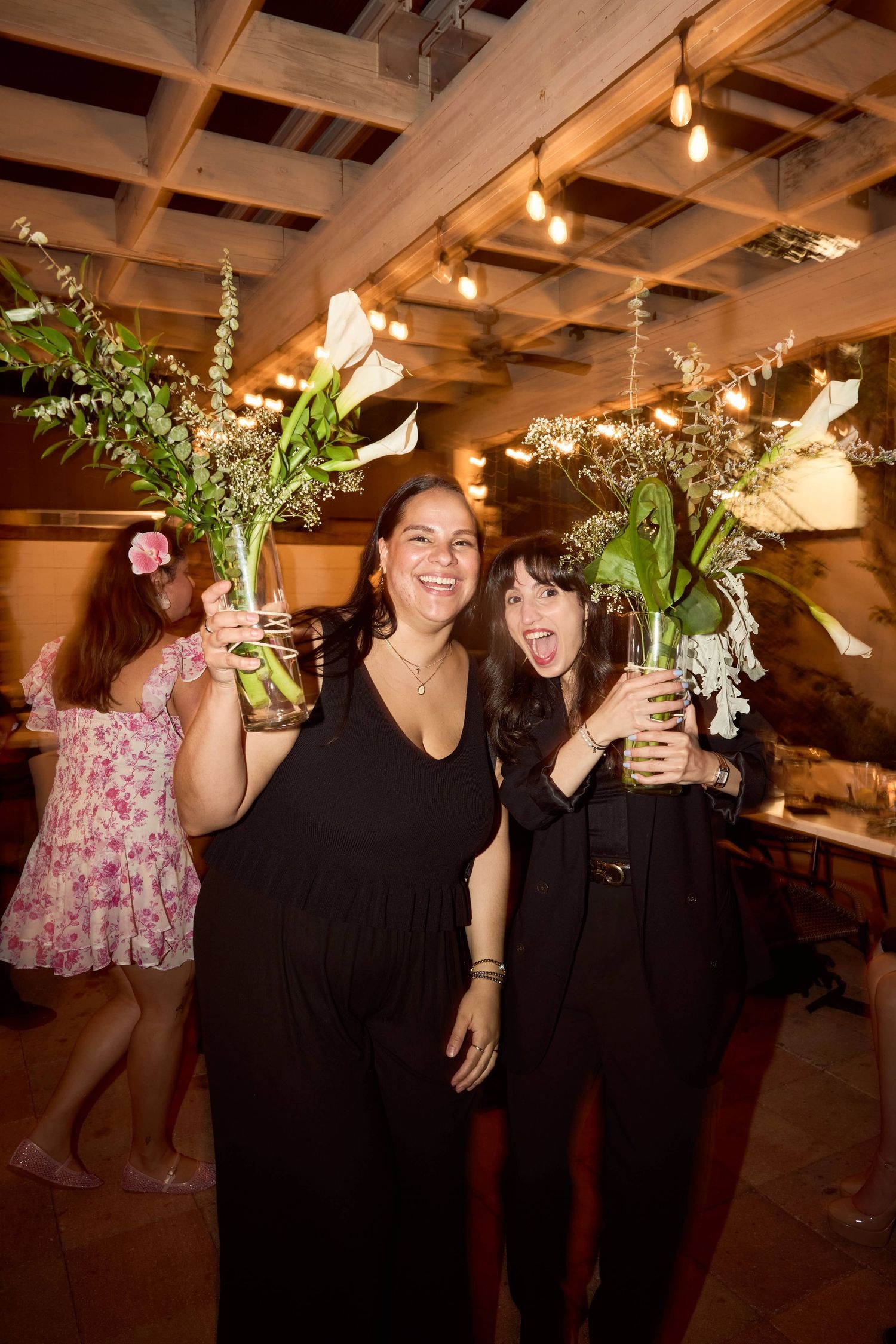 Two women in black dresses smiling and holding large floral arrangements at a warmly lit wedding reception.