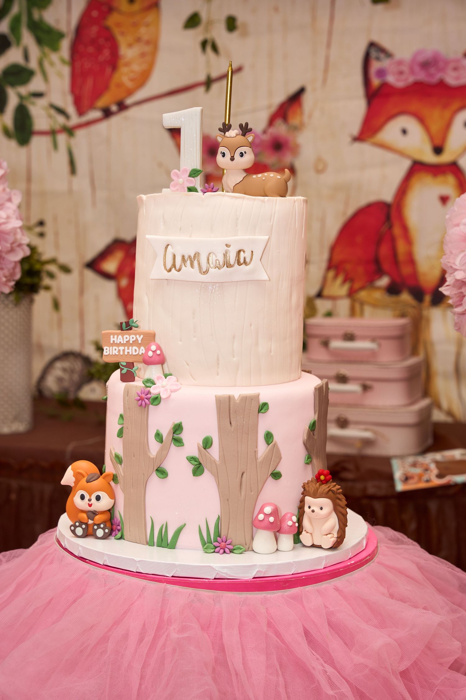 Two-tier woodland themed first birthday cake for Amaia with fox, hedgehog, owl figurines and mushroom decorations.