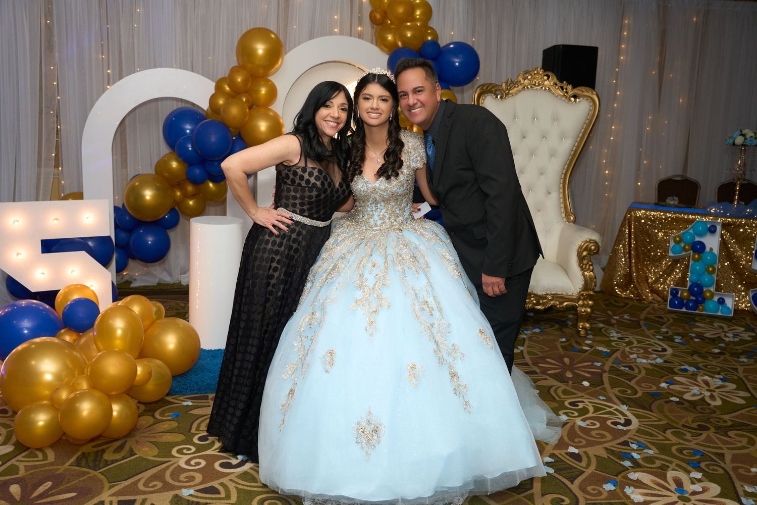 Three people posing at a quinceañera celebration with gold and blue balloon decorations and a white throne chair.