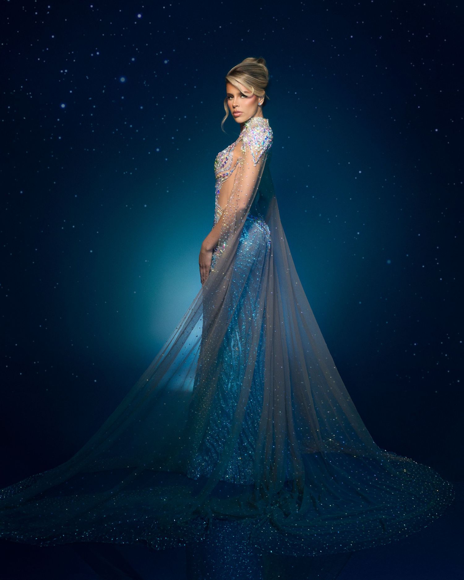 Woman in stunning blue sequined gown with sheer cape poses against starry night sky background.