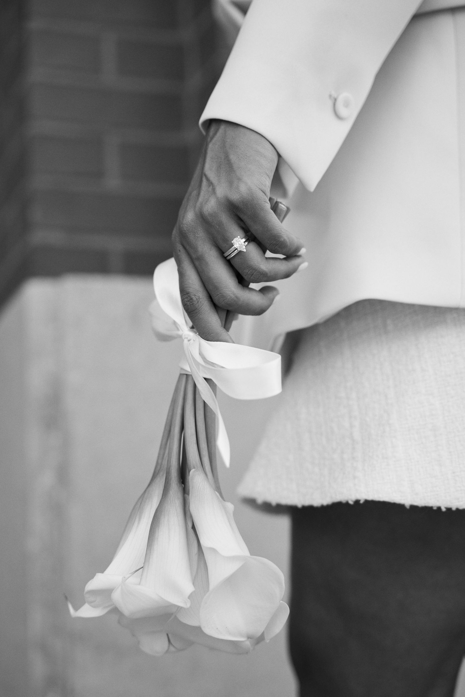 Close-up black and white photo of a hand holding white calla lilies with a ribbon, wearing a diamond ring.