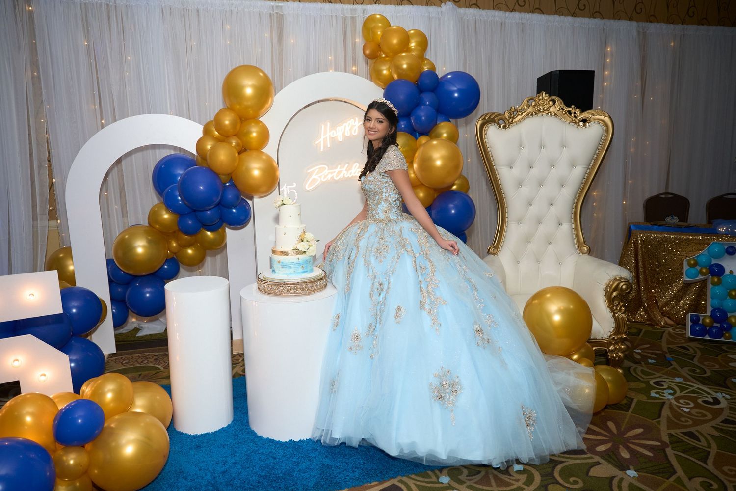 Young girl in light blue quinceañera gown poses with cake, royal blue and gold balloons, and white throne chair.