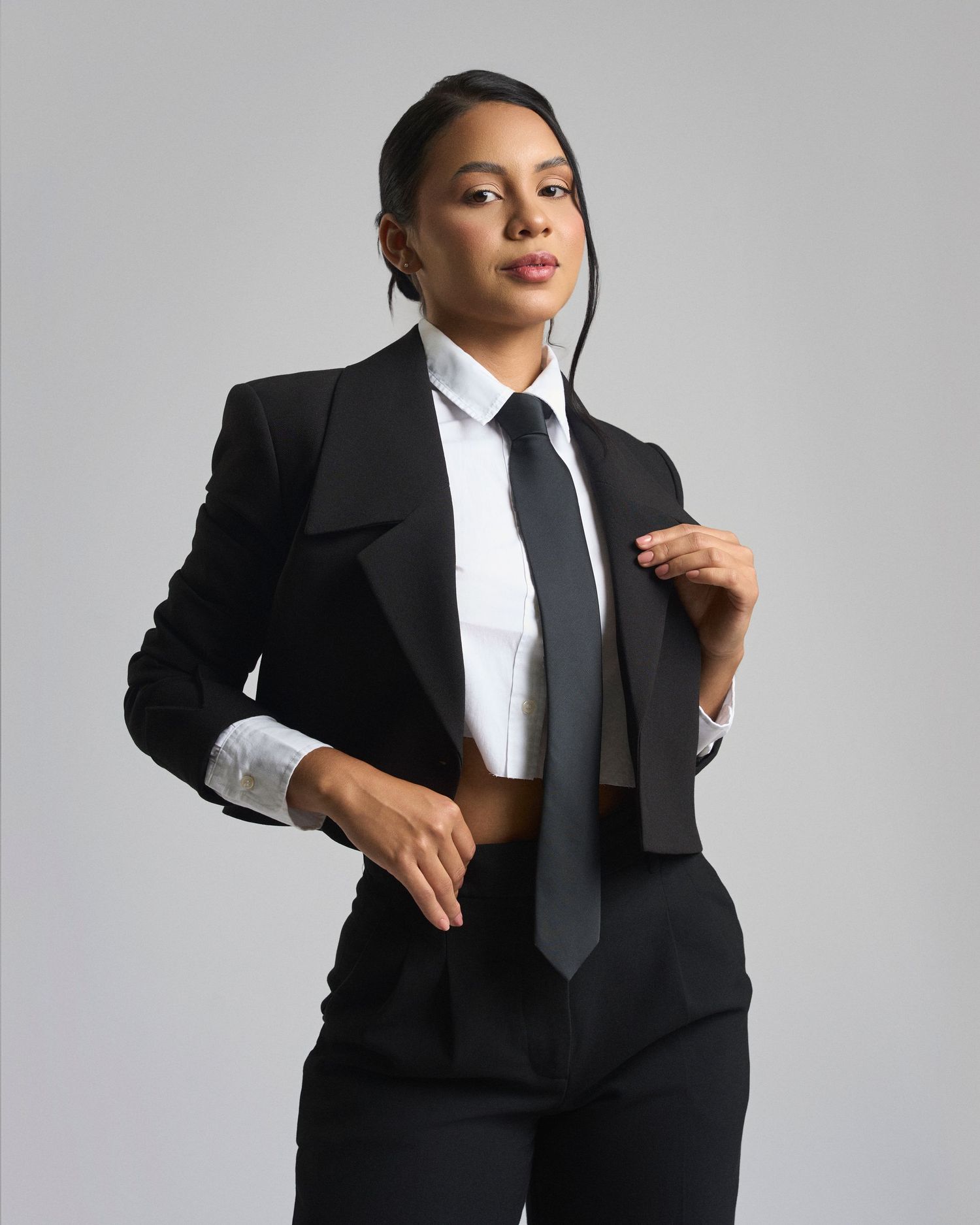 Woman in black tuxedo suit with white shirt and black tie posing against gray background.