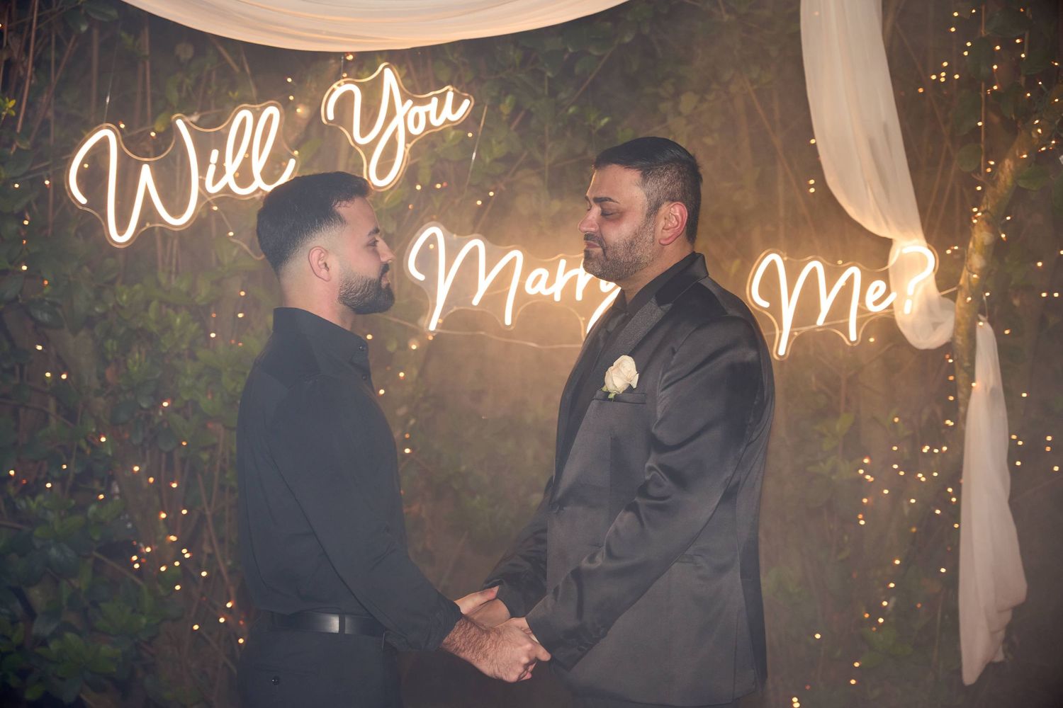 Two men holding hands during a marriage proposal under a neon Will You Marry Me sign with fairy lights.