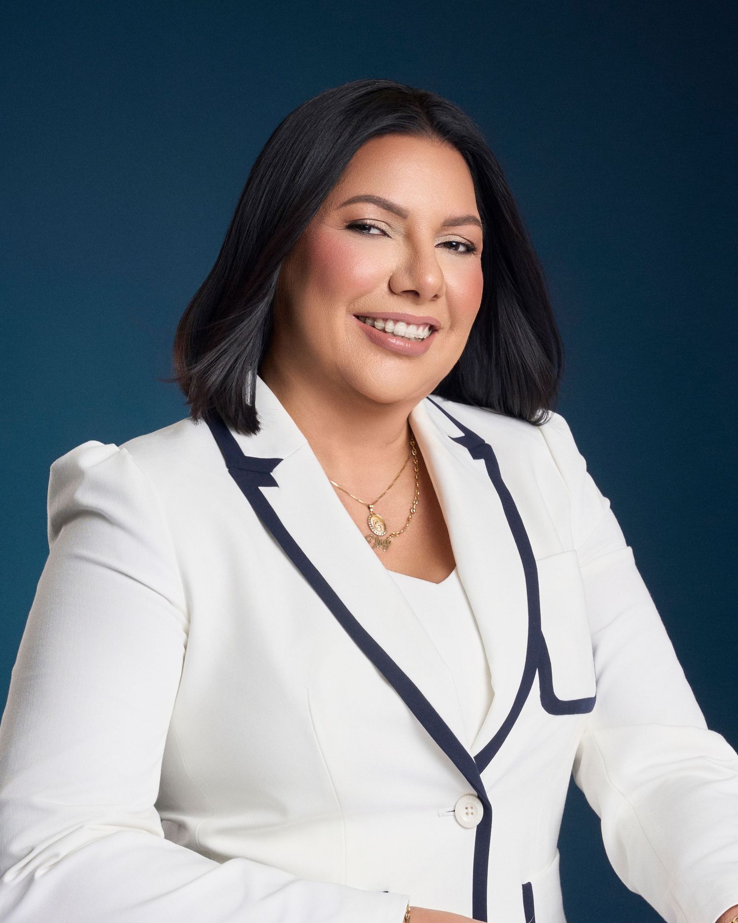 Woman in white blazer with navy trim smiles confidently against dark blue background.