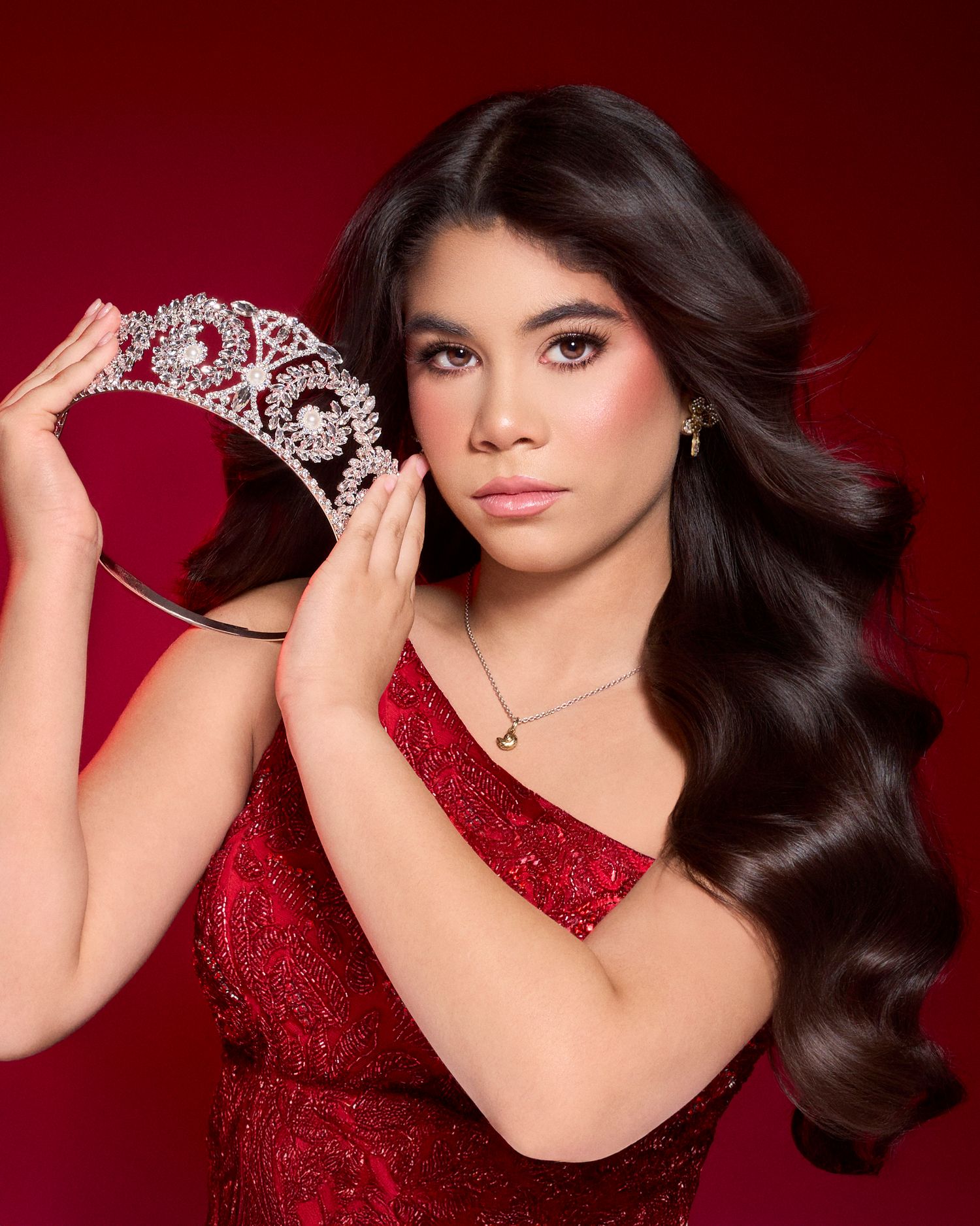 Young woman in red sequined dress holds jeweled crown against vibrant red background.