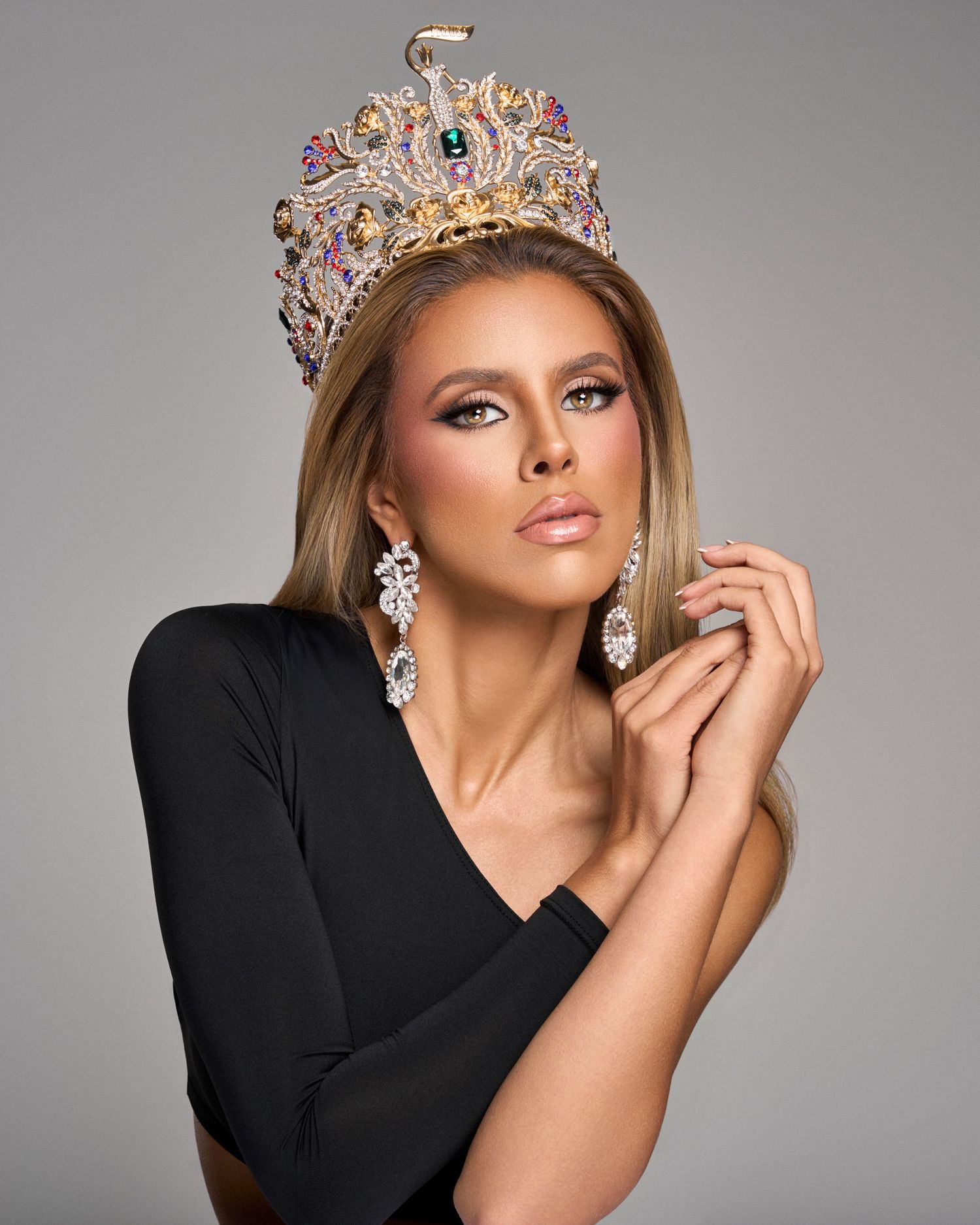 Blonde woman wearing large ornate crown and black dress poses with hands clasped gracefully.