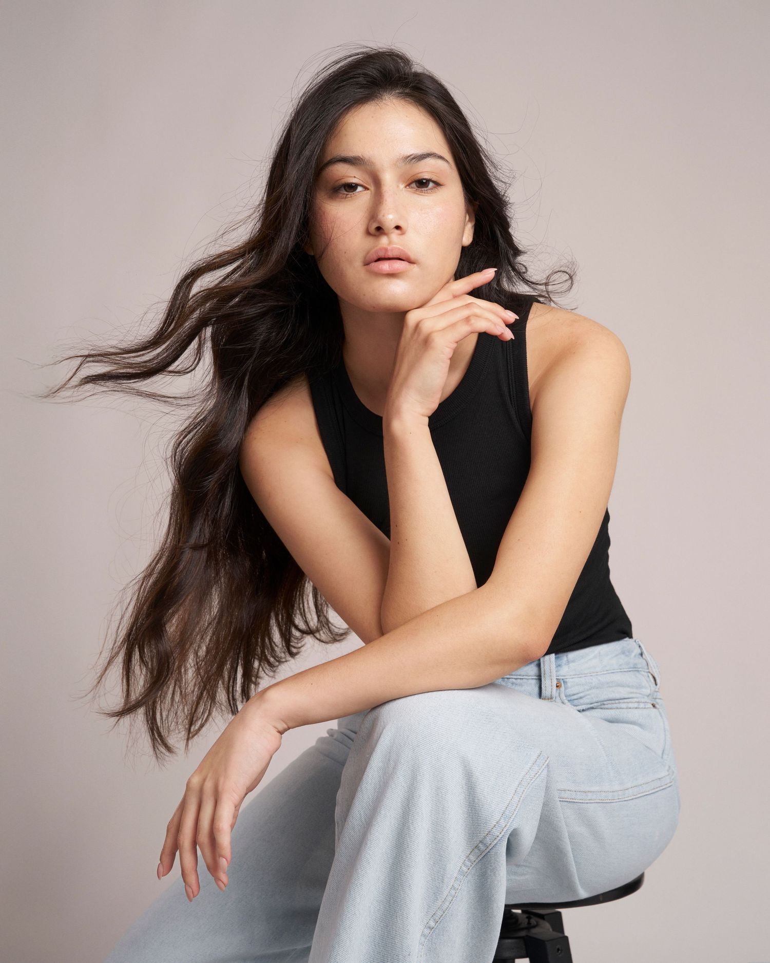 Brunette woman in black sleeveless top and light jeans sitting on stool with hand on chin.