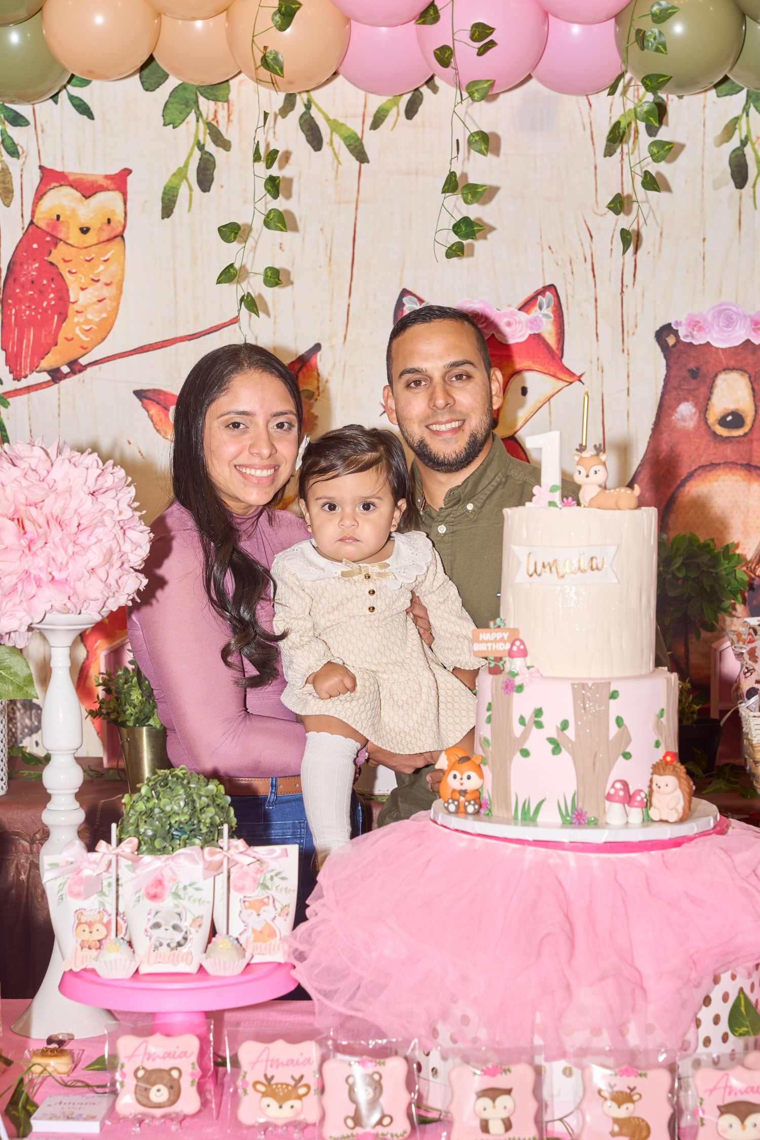 Family celebrating baby girl first birthday party with woodland theme cake, owl decorations, and pink balloons.