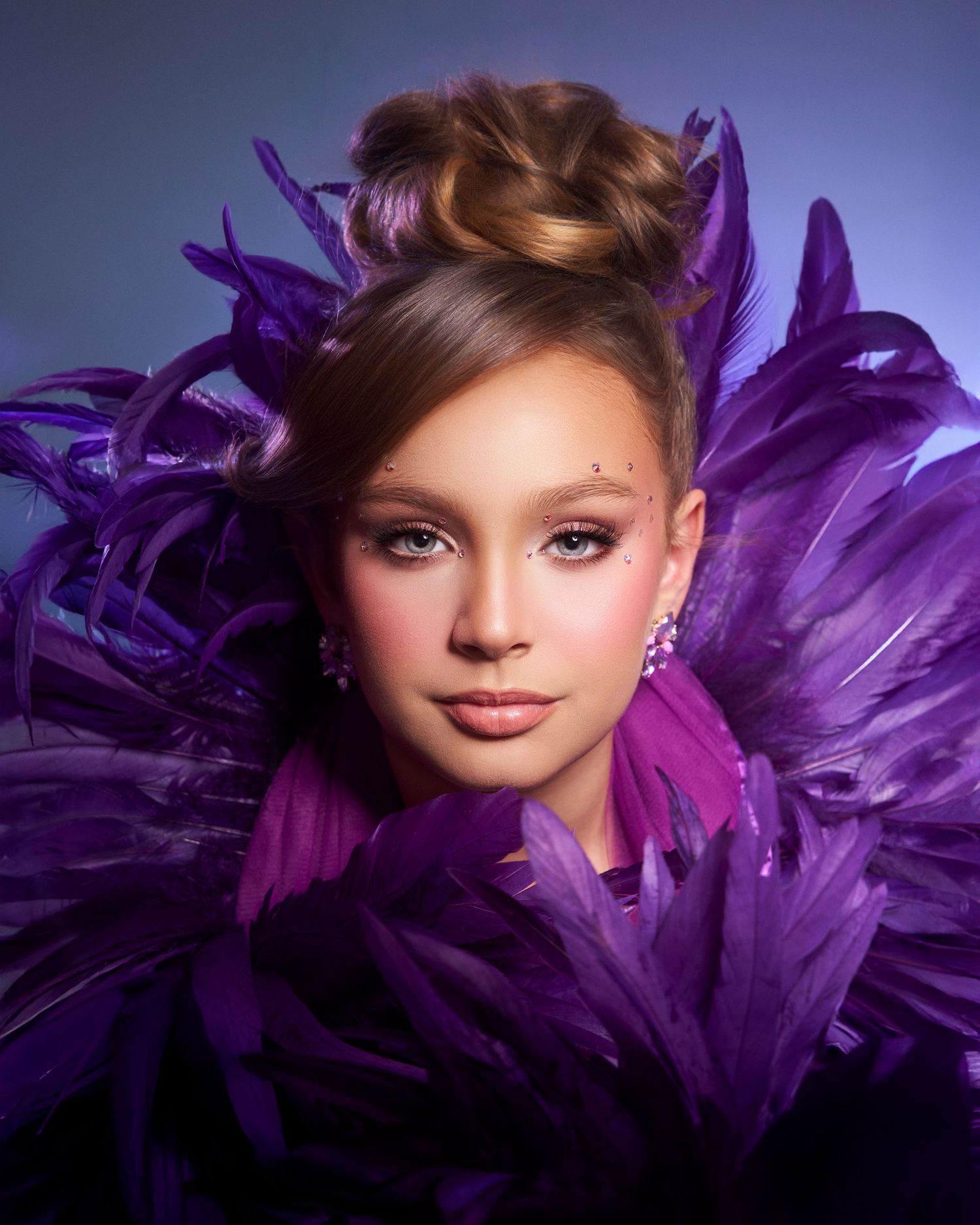 Young woman surrounded by purple feathers with updo hairstyle and glittery eye makeup.