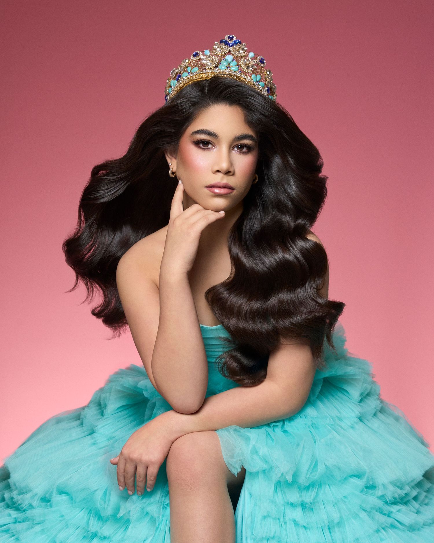 Young beauty queen wearing jeweled crown sits in turquoise ruffled tulle gown on pink background.