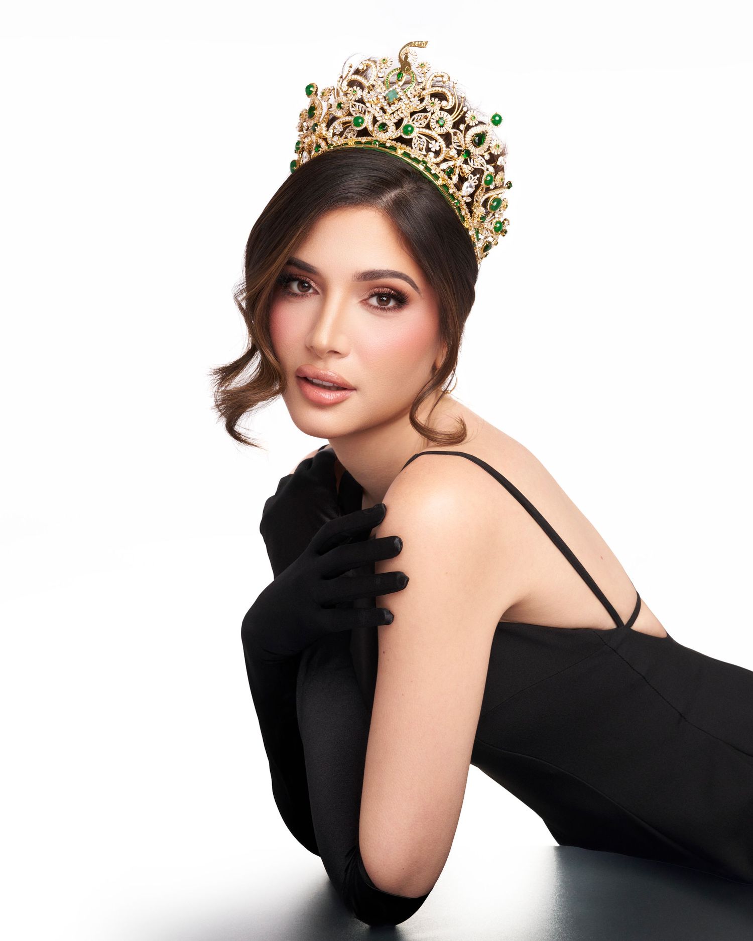 Woman wearing jeweled crown and black gloves poses elegantly against white background.