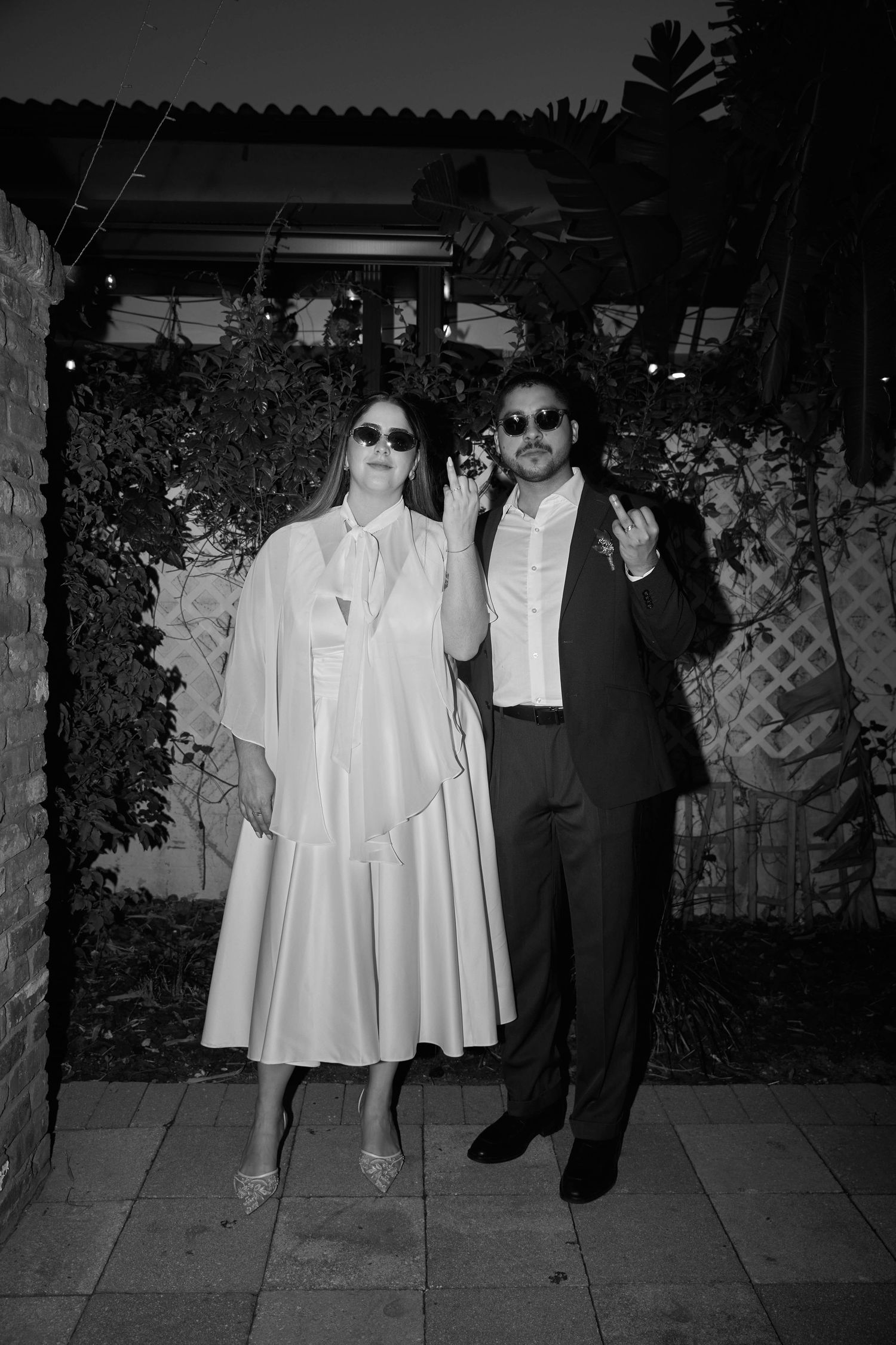 Stylish couple in black and white formal attire posing together at an evening outdoor wedding reception.
