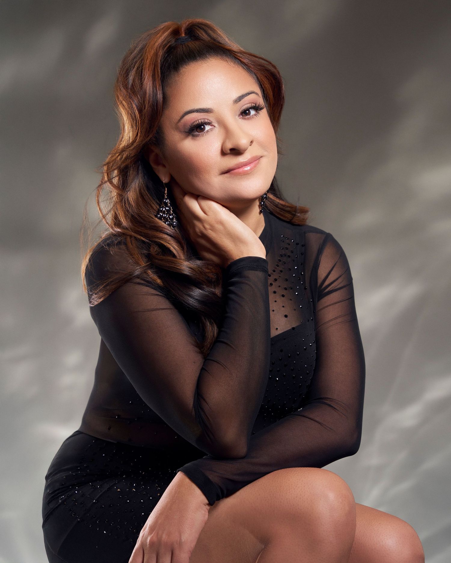 Studio portrait of a smiling woman in a black sheer sparkle dress seated against a gray background.