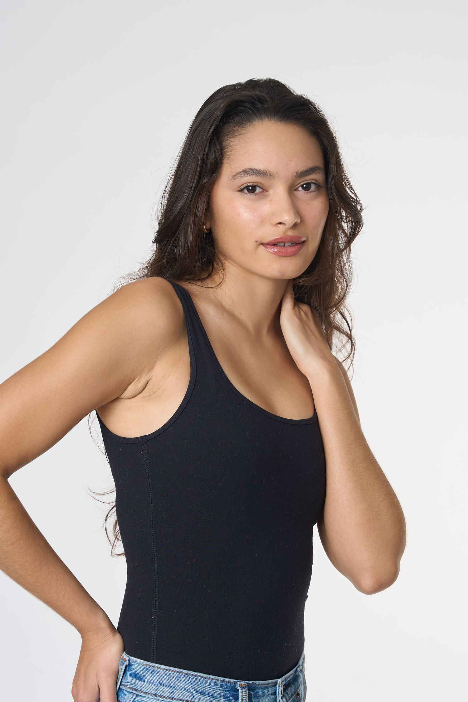 Young woman in black sleeveless bodysuit and jeans poses with hand on neck against white background.