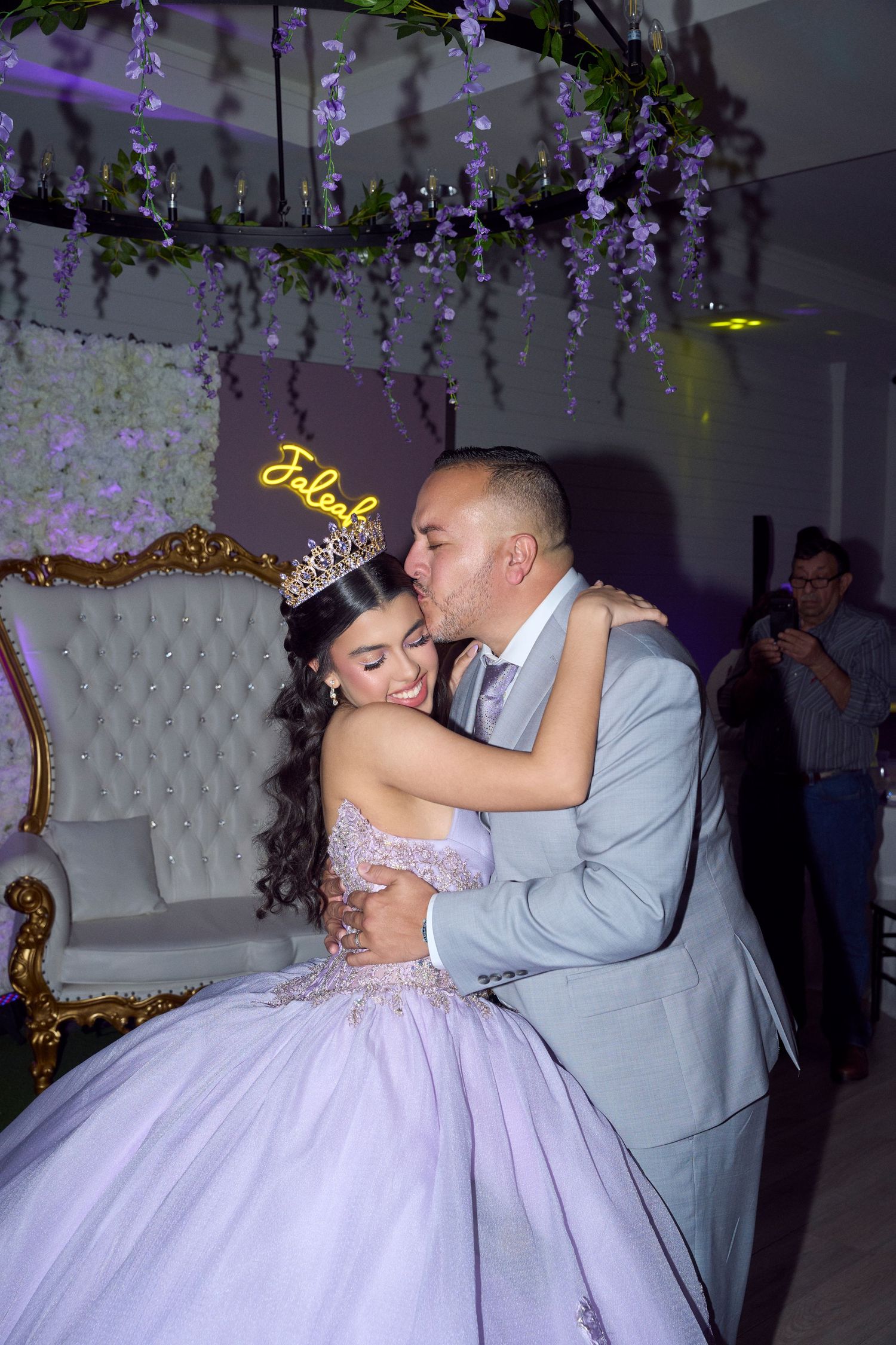 A young woman in a lavender quinceañera gown and tiara shares a tender embrace with a man in a gray suit at her celebration.