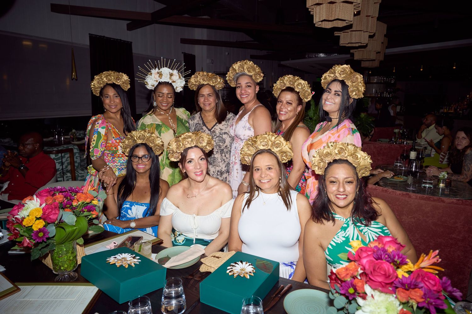 Group of women in tropical floral outfits and golden headdresses celebrating at a festive dinner party with colorful flower arrangements.