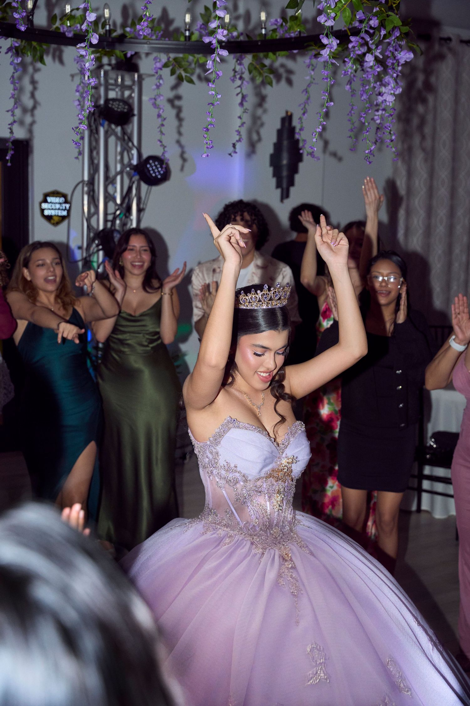Young woman in lavender quinceañera ball gown and tiara dancing with raised arms at festive celebration.