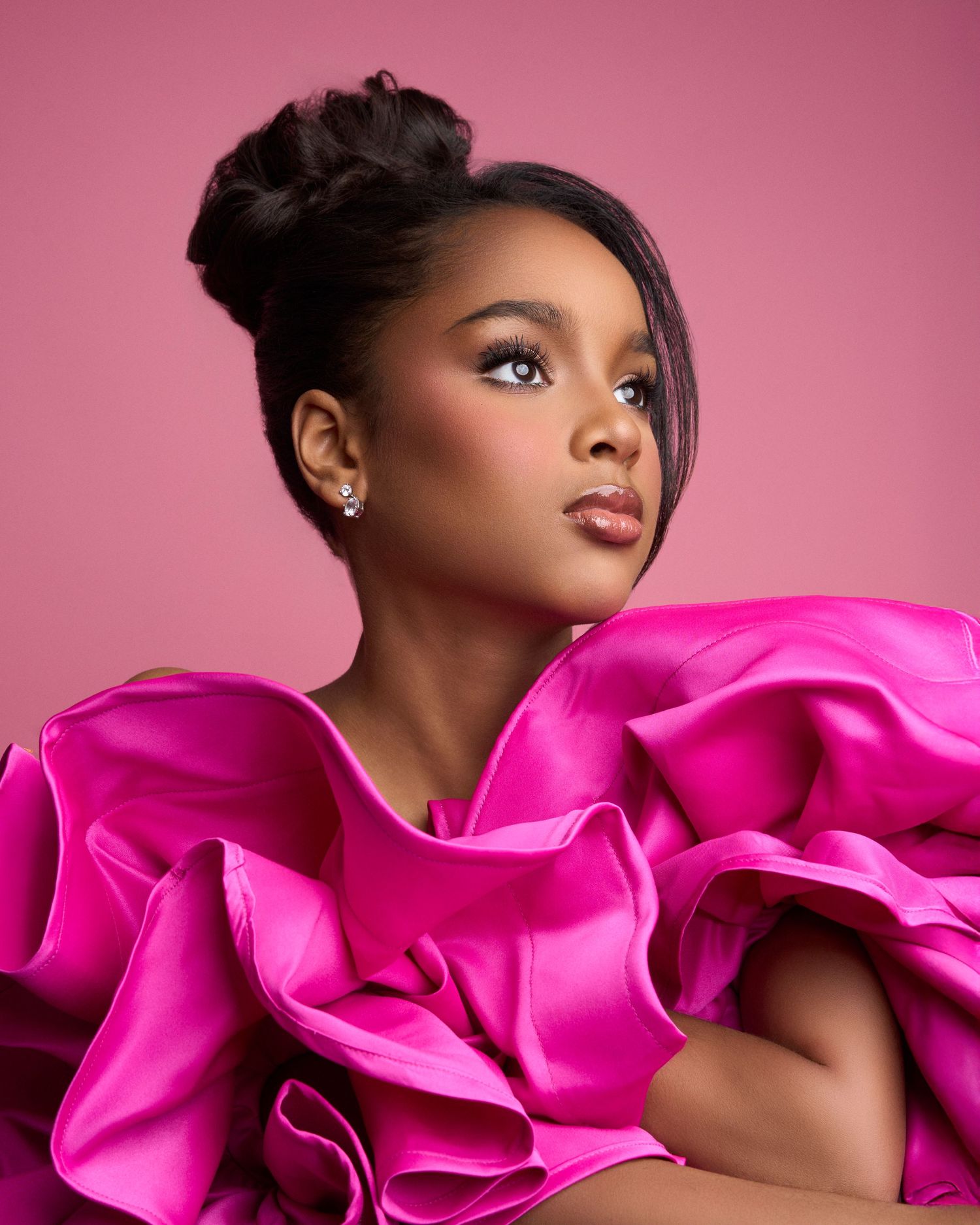 Young girl in dramatic pink ruffled satin dress poses elegantly against pink background.