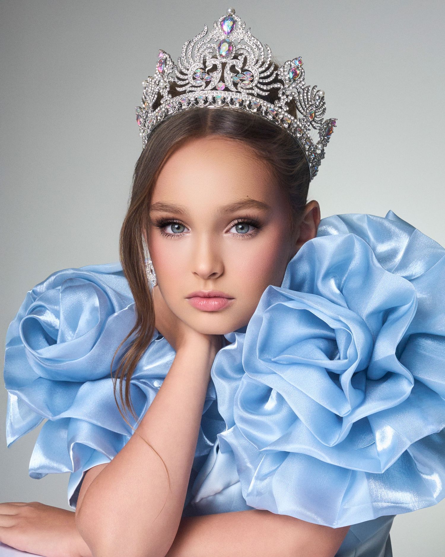 Young girl wearing a large silver rhinestone crown and blue ruffled satin dress in pageant portrait.
