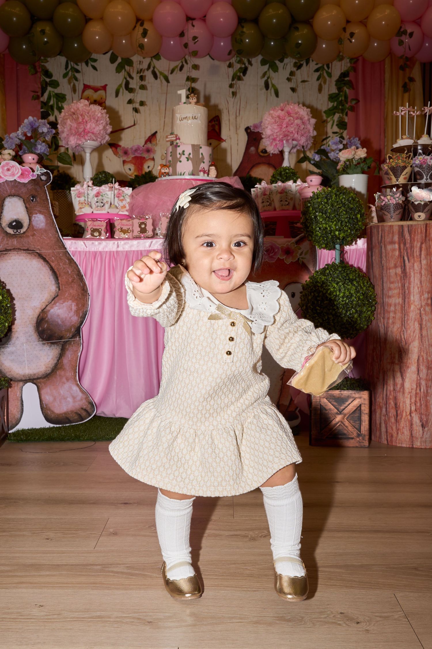 Toddler girl in cream dress and gold shoes dancing at a pink woodland-themed first birthday party.